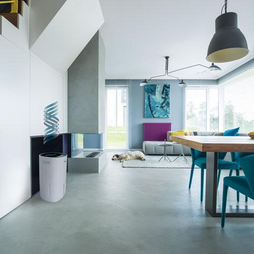Ein weißer Luftreiniger steht neben einem modernen Kamin in einem Wohnzimmer mit einem Hund auf dem Teppich.