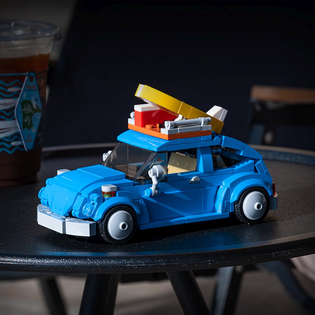 Niebieski samochód z klocków Lego z czerwoną lodówką i żółtą deską surfingową na dachu.