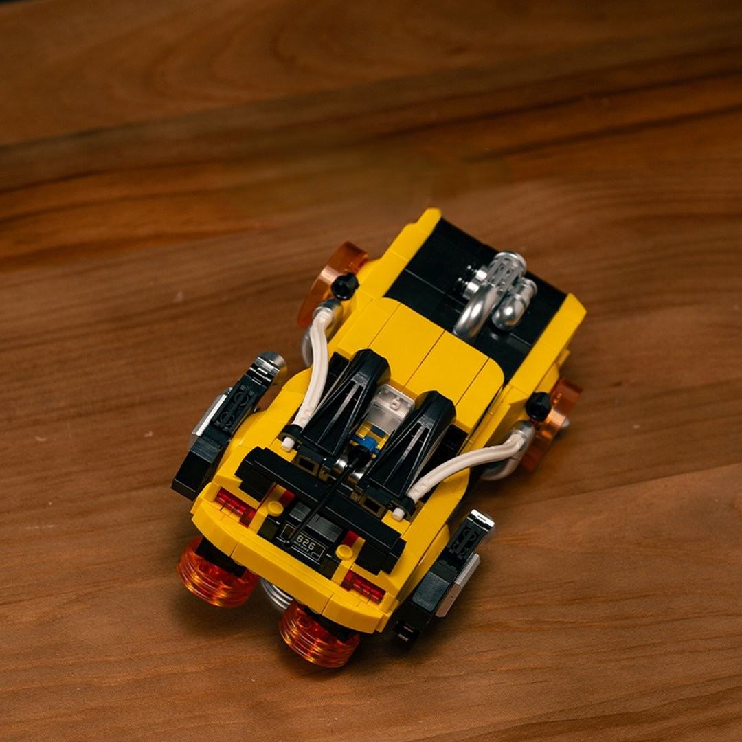 Żółty samochód Lego z czarnymi akcentami i pomarańczowymi kołami na drewnie.