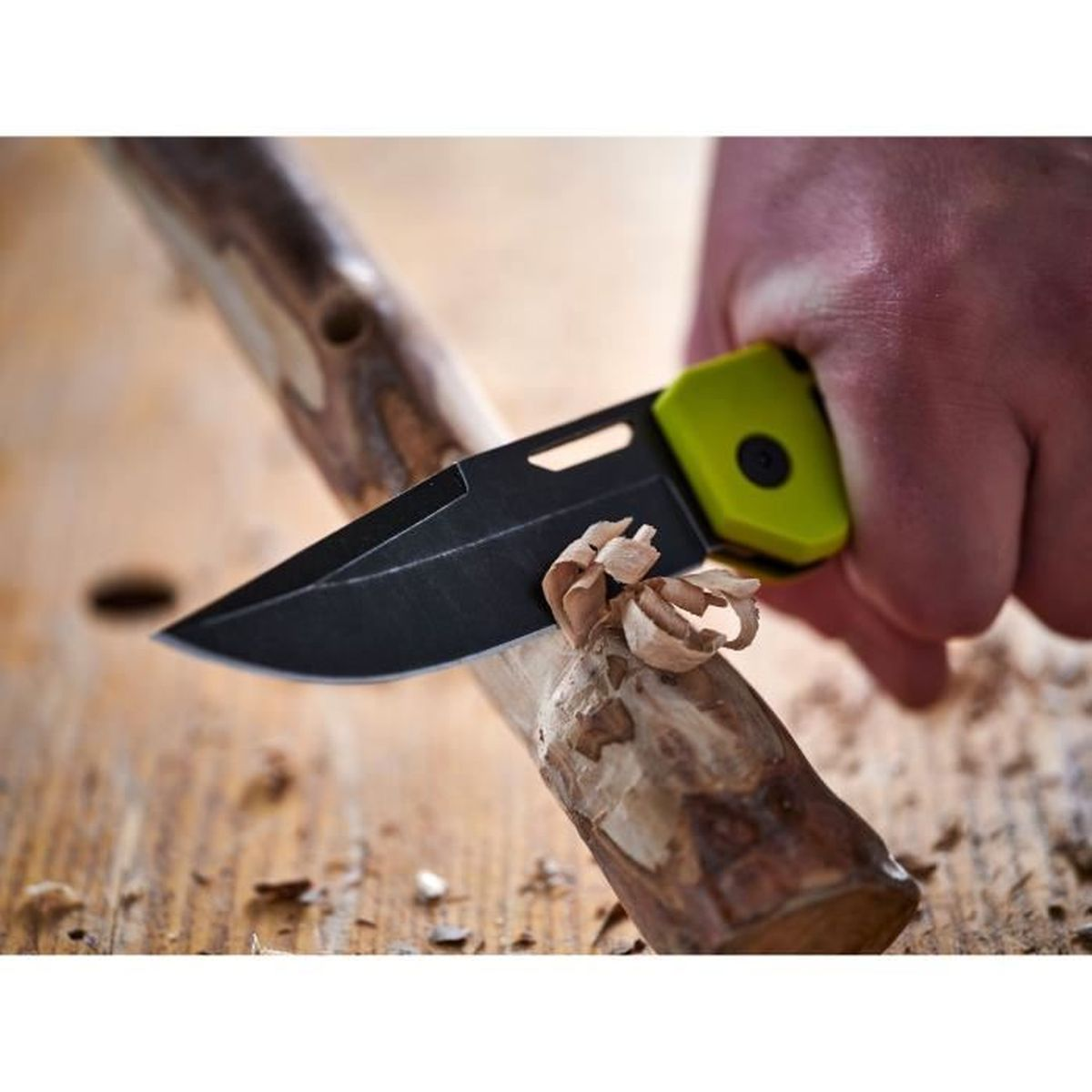 Un cuchillo de hoja negra y mango verde neón talla virutas de madera de una rama.