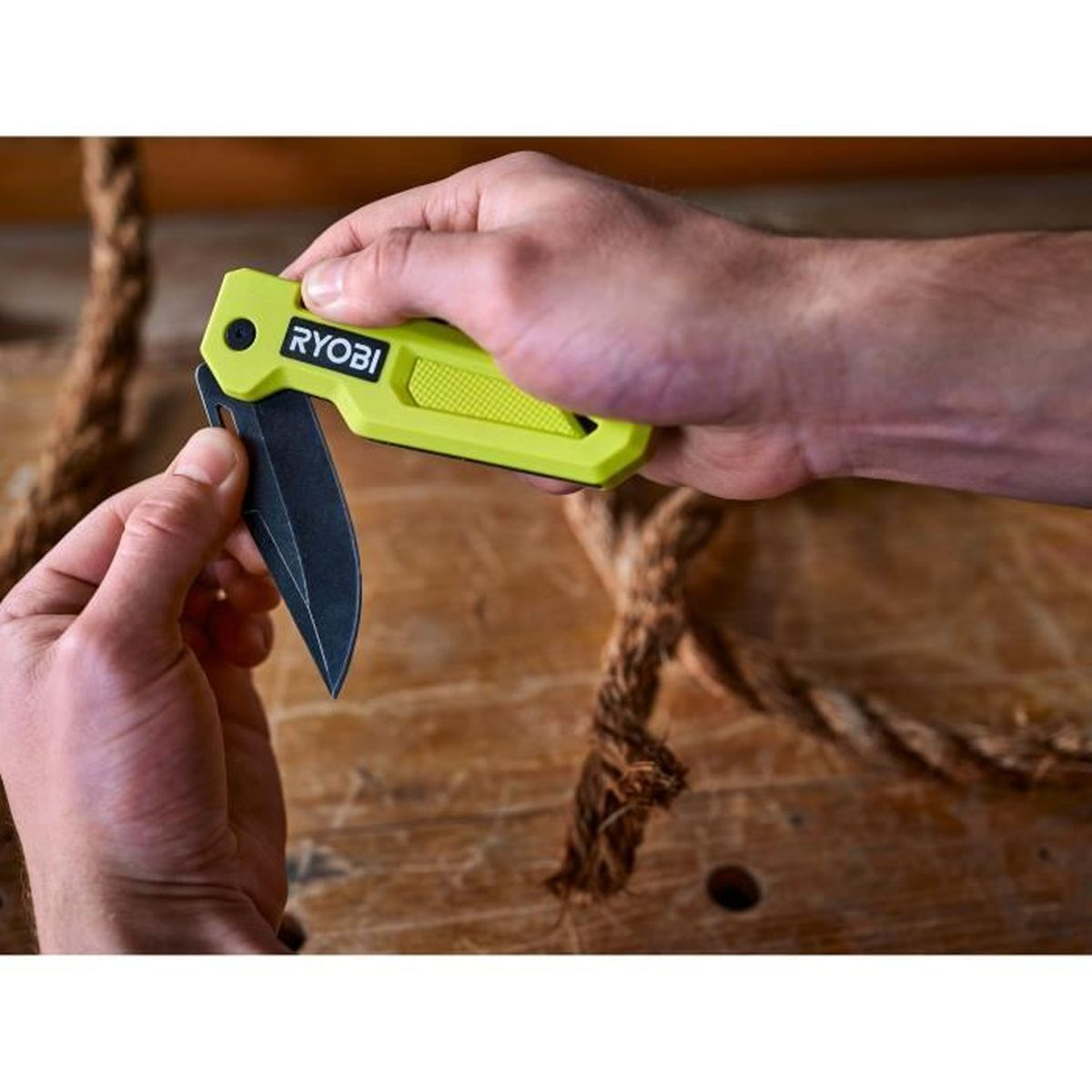 Una mano sostiene un cuchillo Ryobi amarillo con hoja negra sobre una cuerda.