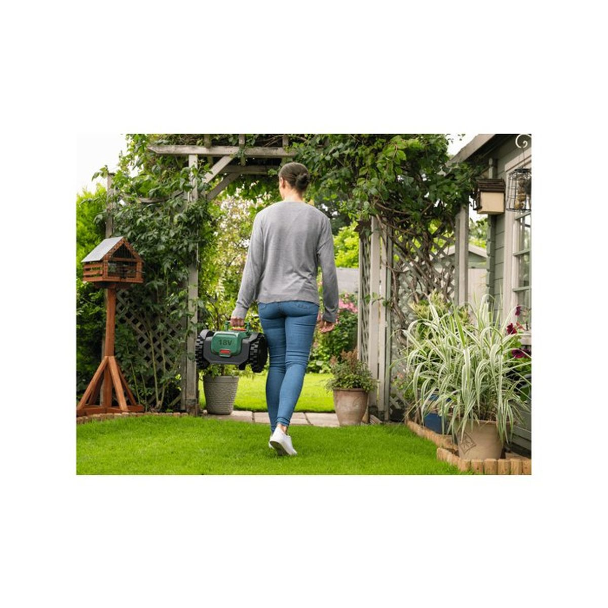 Eine Frau trägt einen grünen Rasenmähroboter durch einen Garten mit Bäumen und Pflanzen.