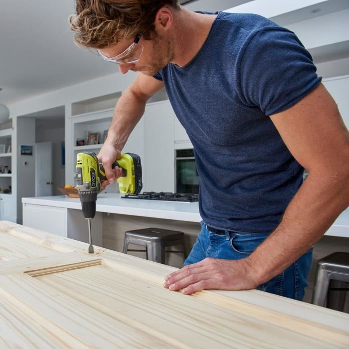 Hombre con camisa azul y jeans taladra tabla de madera en cocina moderna y luminosa.