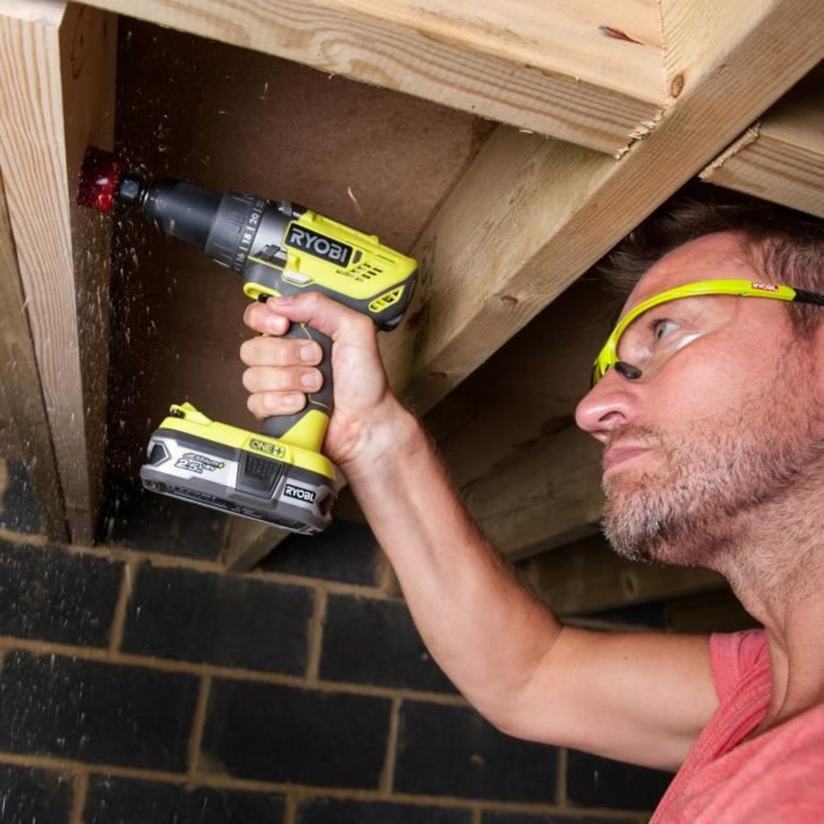 Hombre taladra agujero en madera con taladro amarillo, lleva gafas.