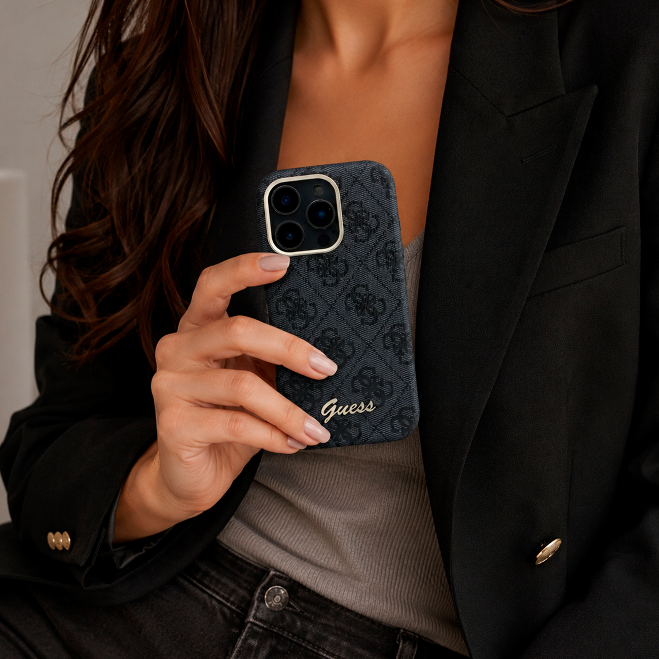 Una mujer sostiene una funda de teléfono gris oscuro con logo y patrón Guess.