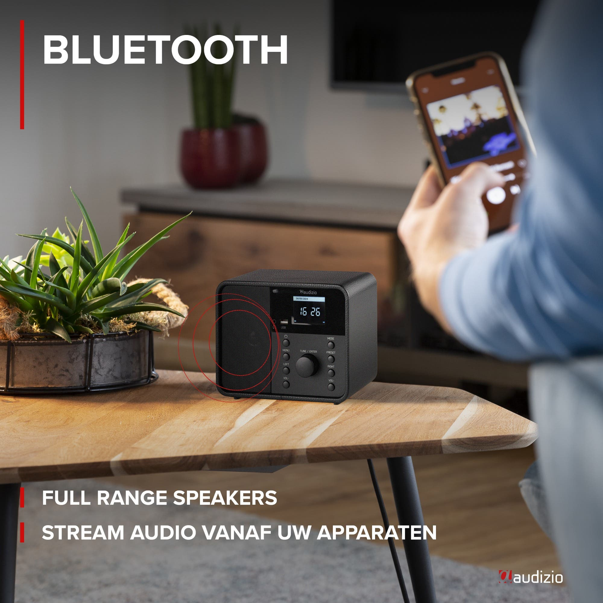 Ein schwarzer Audizio Bluetooth-Lautsprecher steht auf einem Holztisch, daneben eine Pflanze. Eine Person hält ein Smartphone.