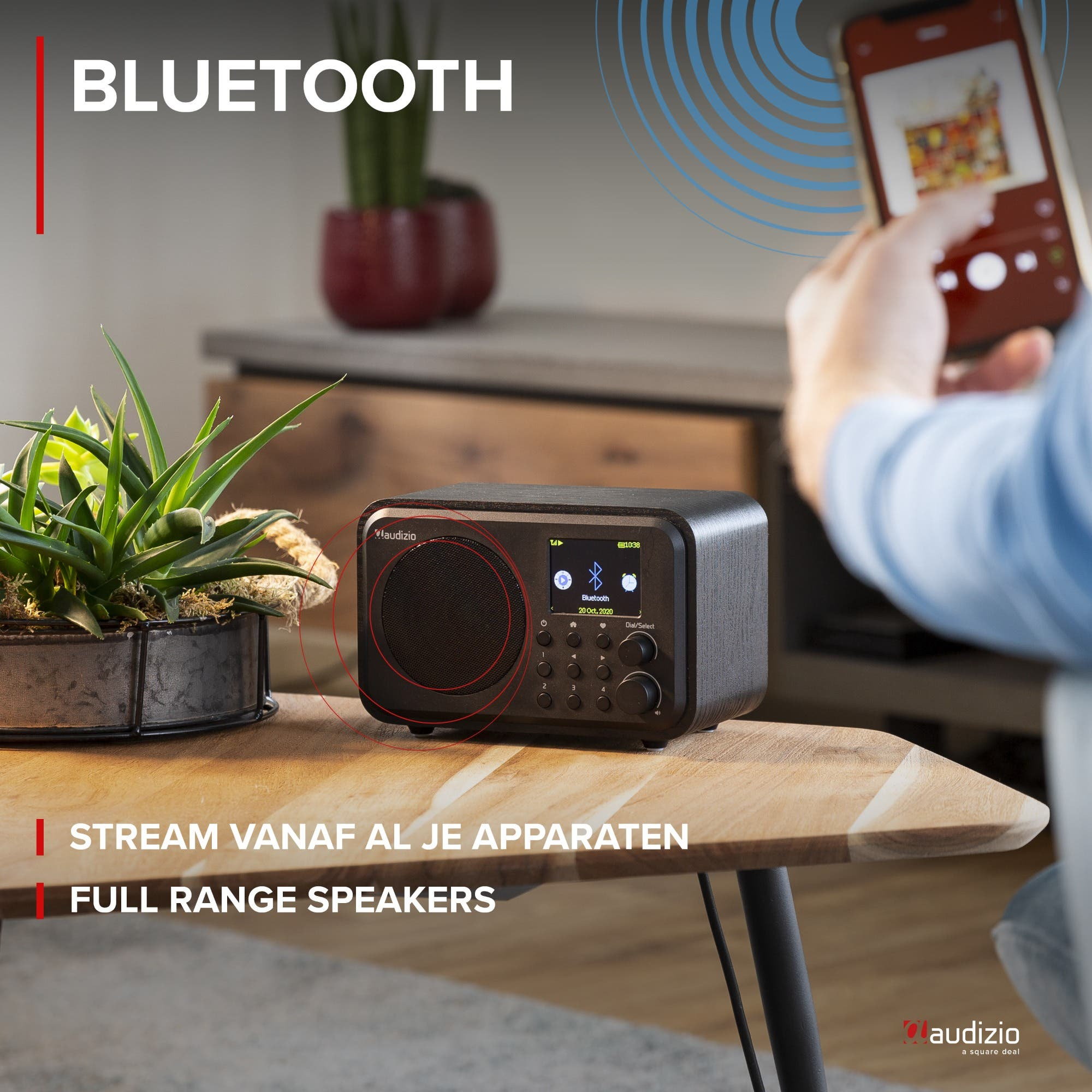 Una radio Bluetooth Audizio con plantas sobre una mesa de madera. Una mano sostiene un smartphone.