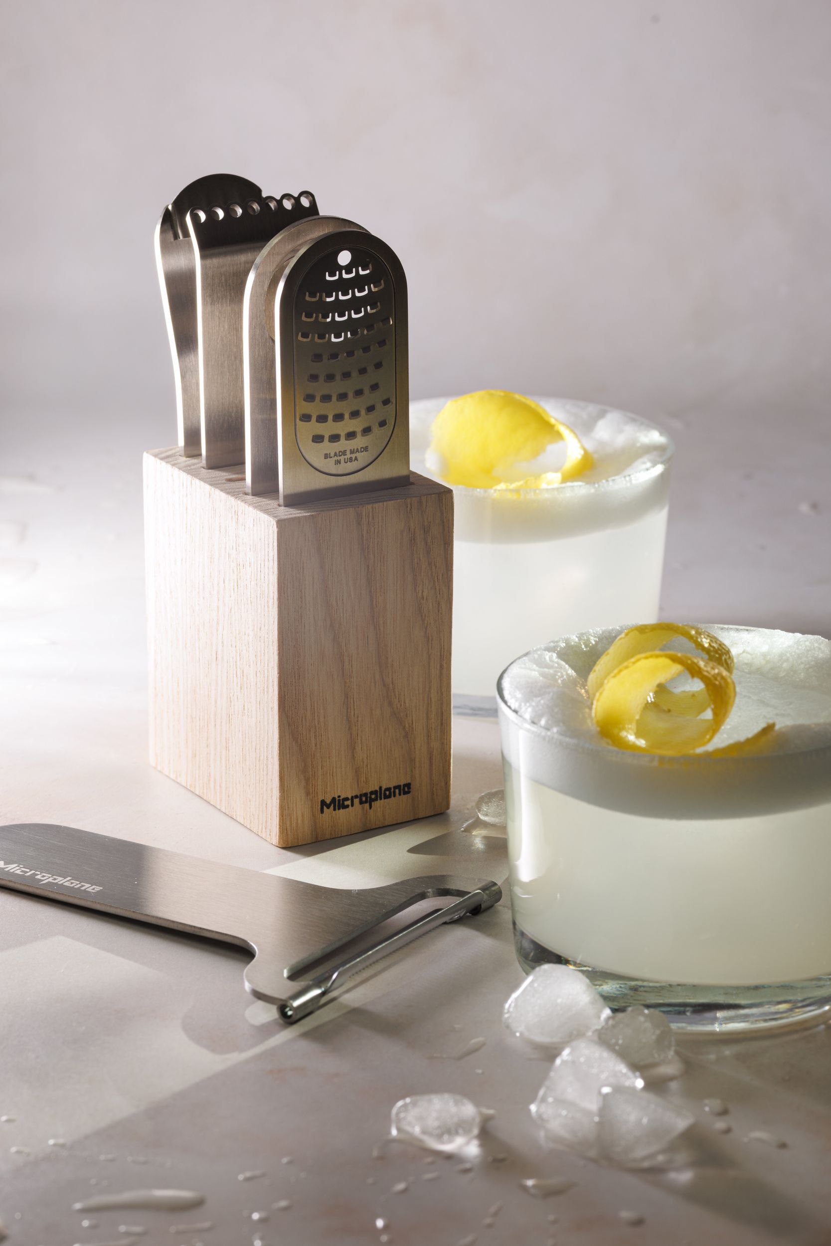 Ein Holzmesserblock mit drei Reiben, zwei Cocktails mit Zitronenzeste und Eiswürfeln.