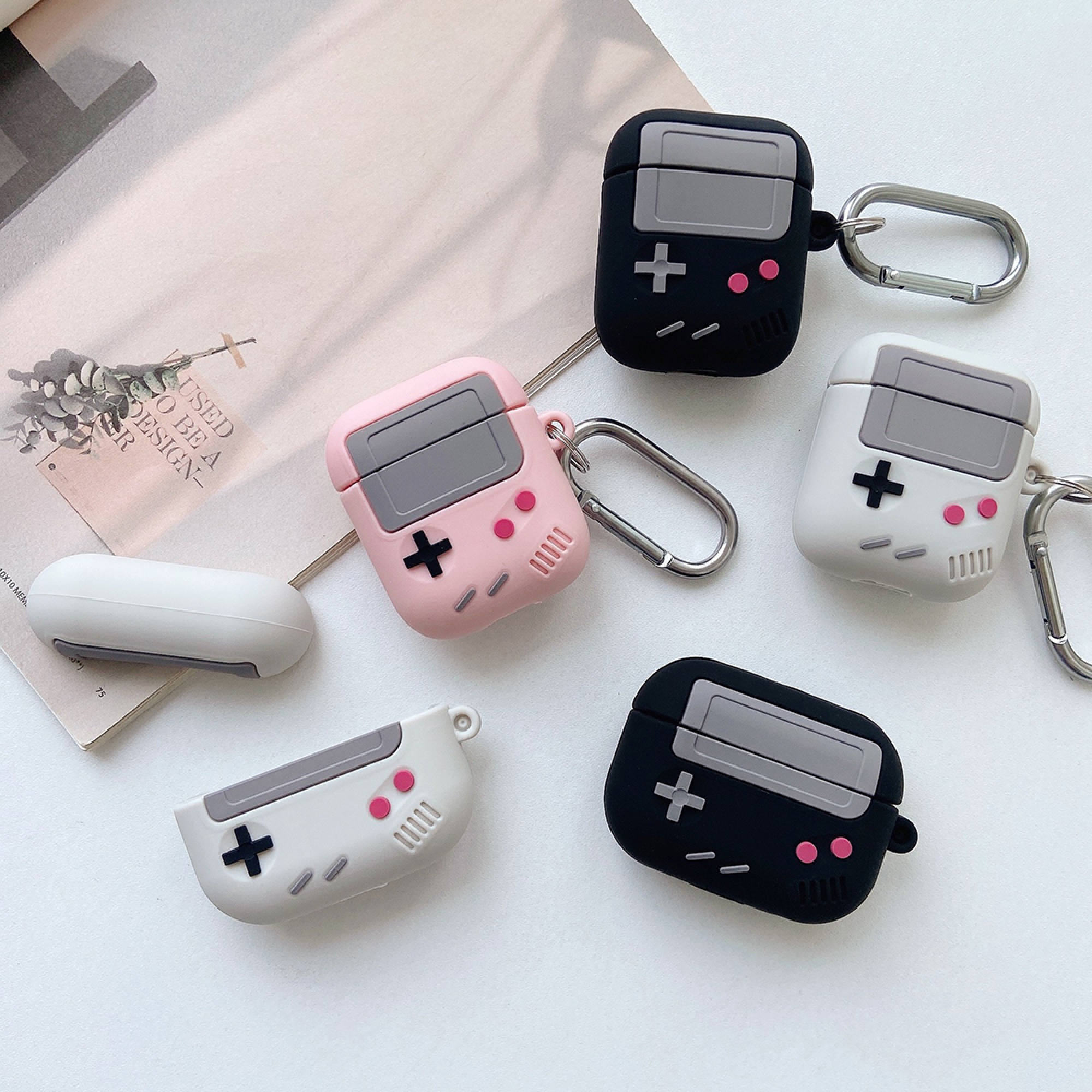 Fünf AirPod-Hüllen im Stil von Handheld-Spielkonsolen, zwei schwarz, zwei weiß, eine rosa.