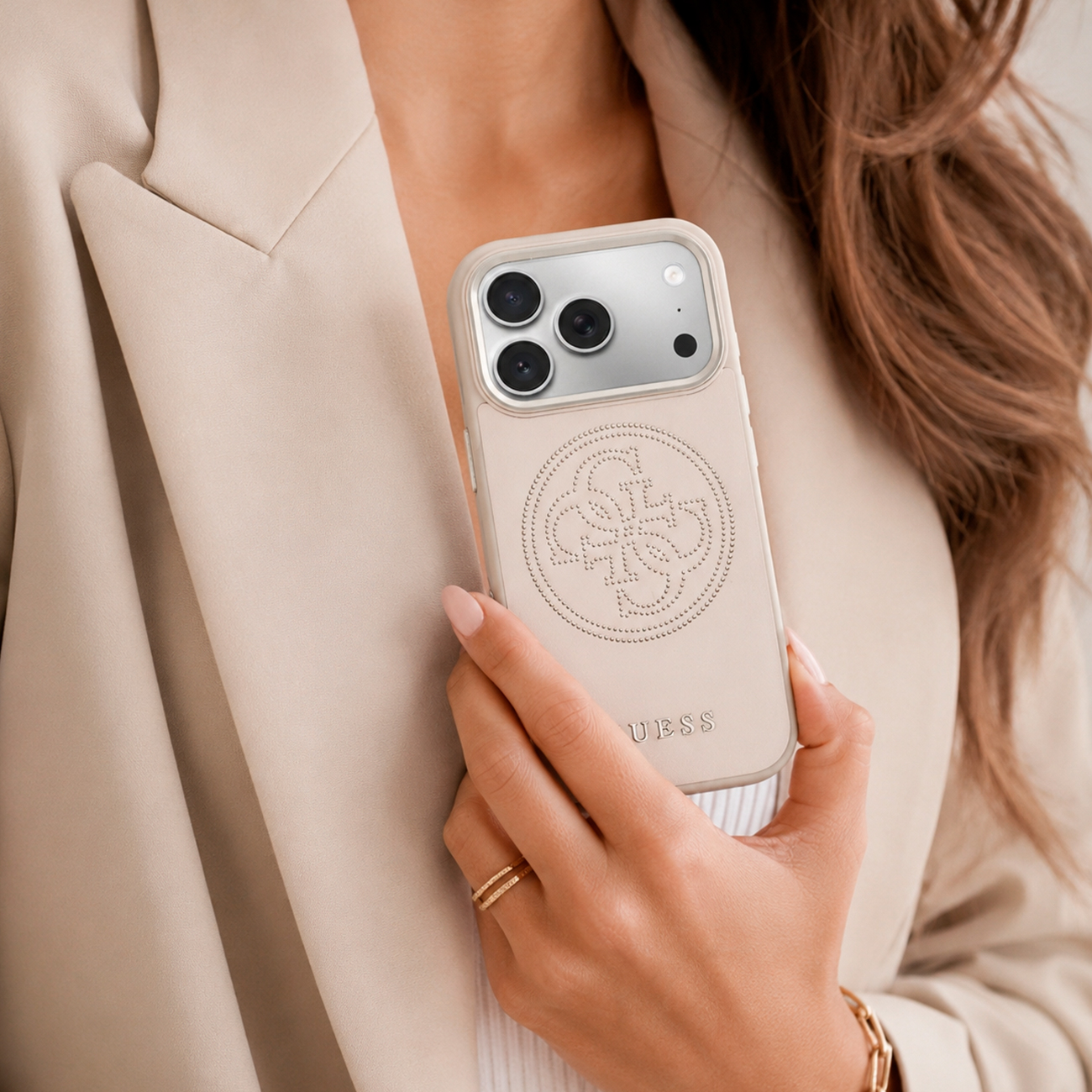 Una donna tiene un telefono beige chiaro con logo Guess davanti a un blazer beige.