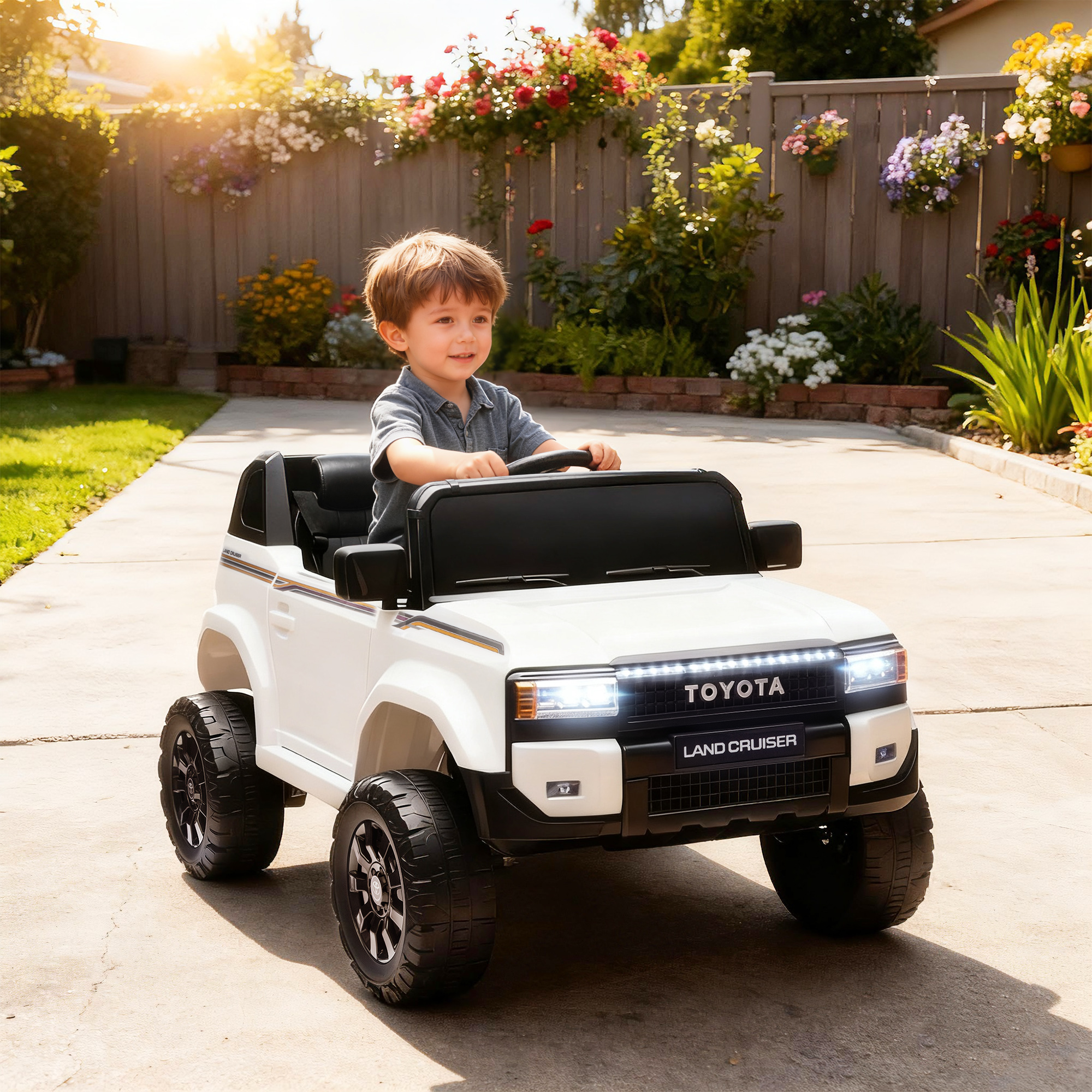 Un bambino guida un Land Cruiser giocattolo bianco in un cortile soleggiato con fiori.