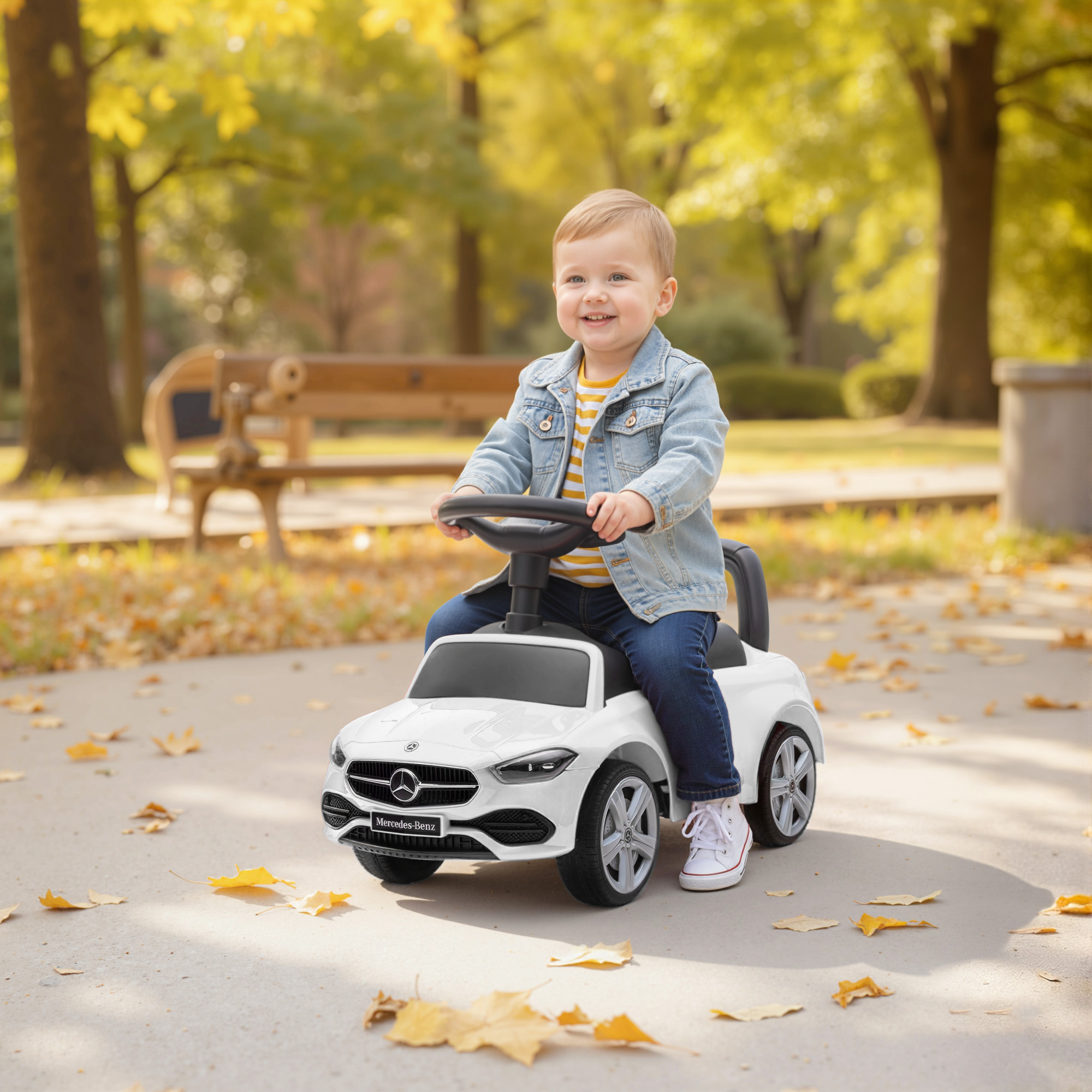 Un bimbo sorridente in giacca di jeans guida un'auto giocattolo Mercedes bianca in un parco.