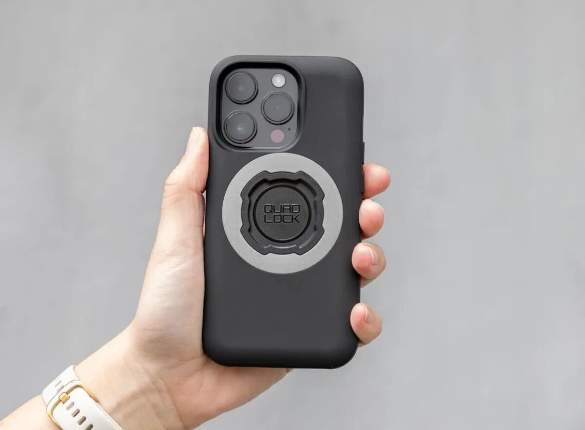 Hand hält schwarzes Handy mit Quad Lock Halterung. Grauer Hintergrund.
