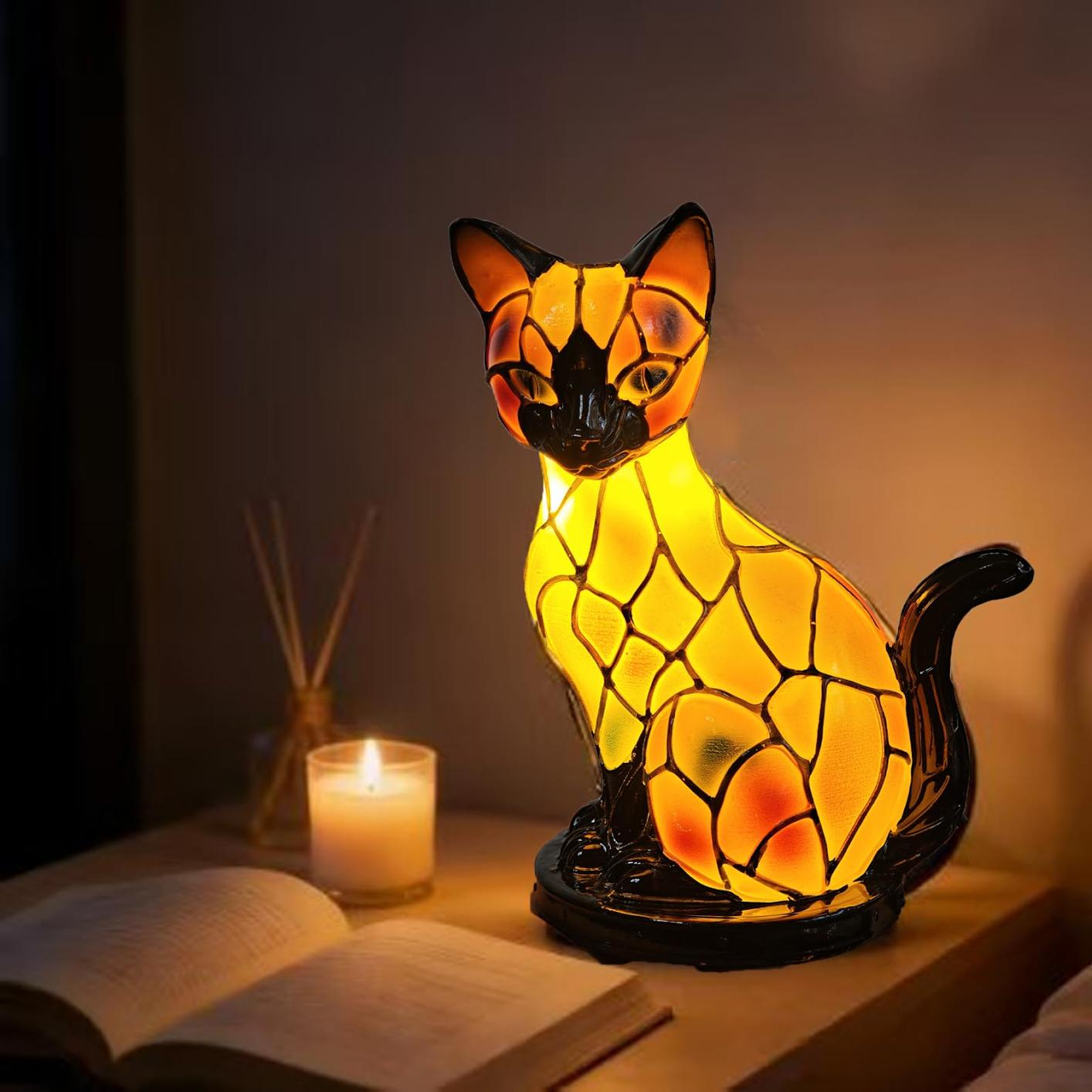 Eine Tiffany-Katzenlampe leuchtet warm auf einem Nachttisch neben einer brennenden Kerze und einem offenen Buch.