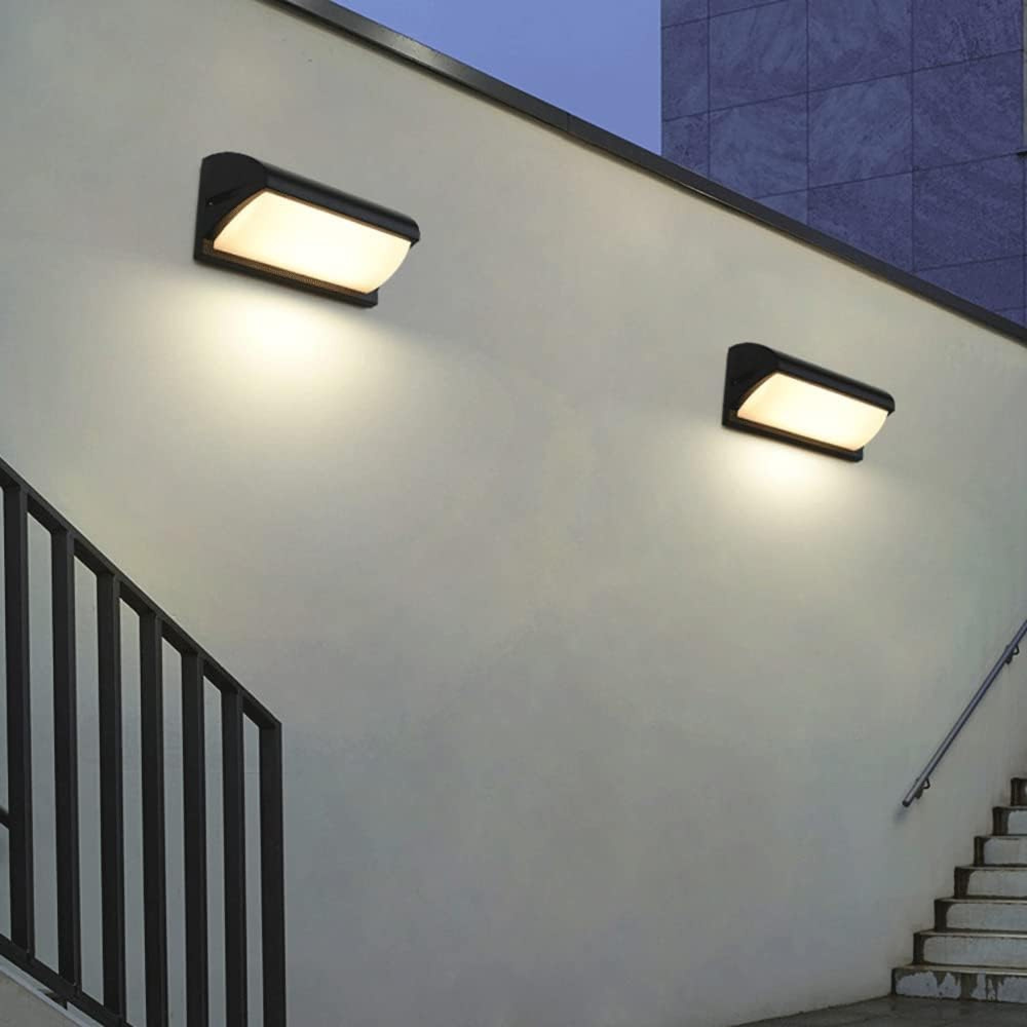 Zwei Wandleuchten beleuchten eine weiße Wand neben einer Treppe und einem Geländer.