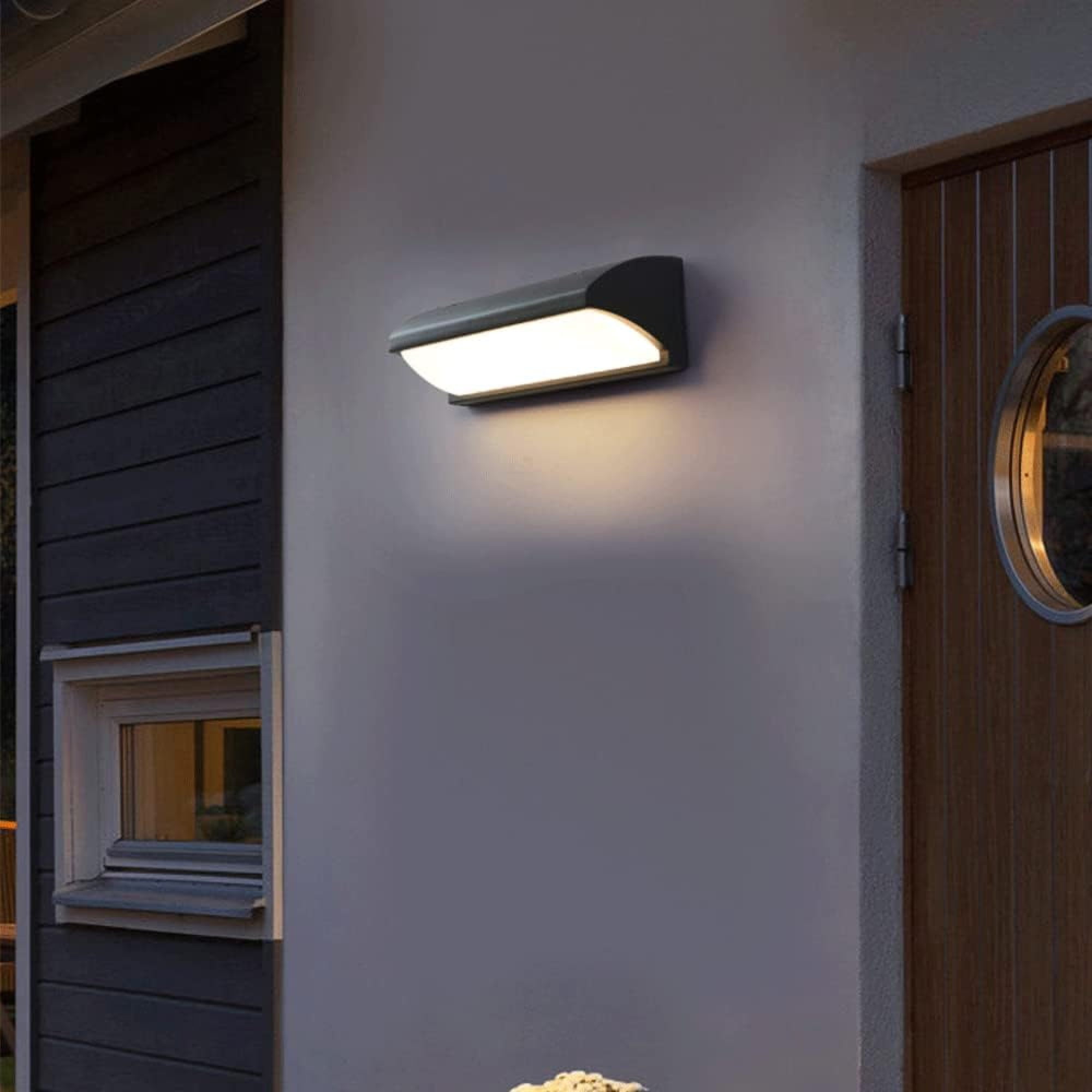 Eine dunkle Wandleuchte beleuchtet eine graue Wand neben einer Holztür.