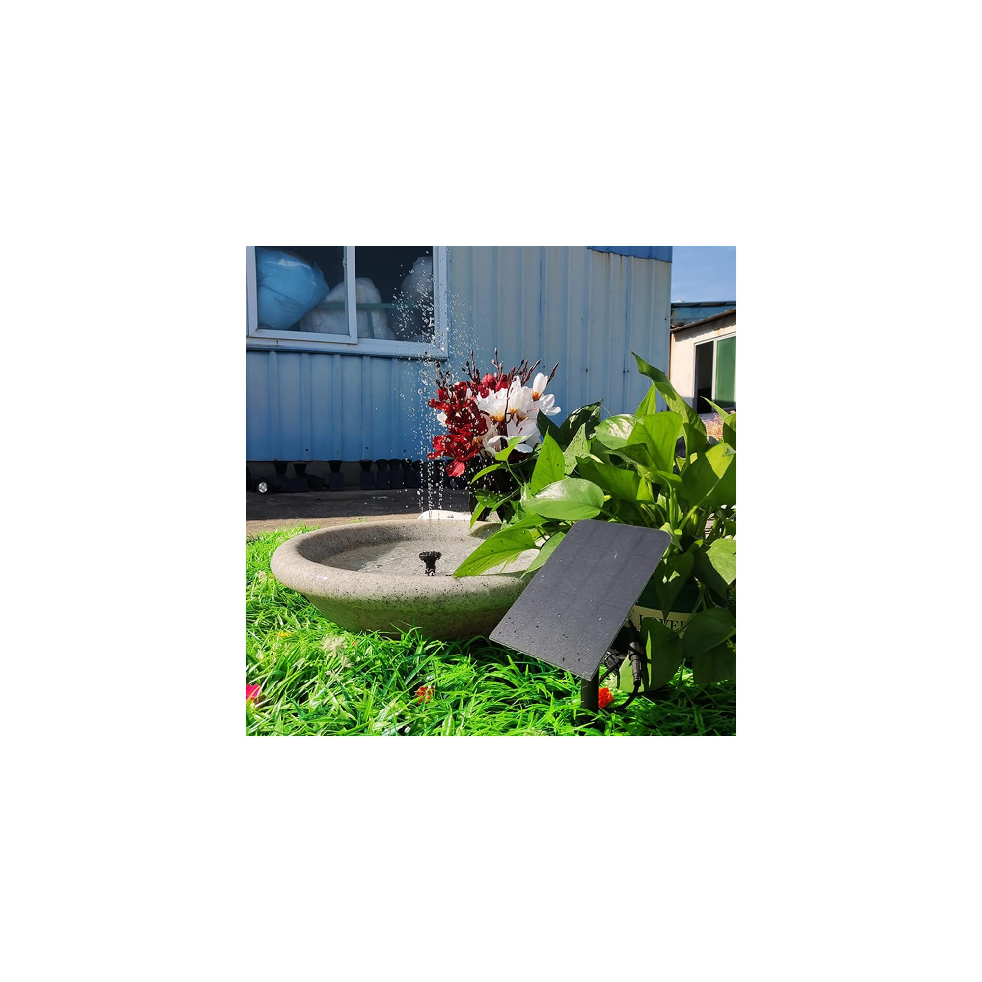 Ein Solar-Springbrunnen mit Blumen und Pflanzen auf Rasen vor einem blauen Gebäude.