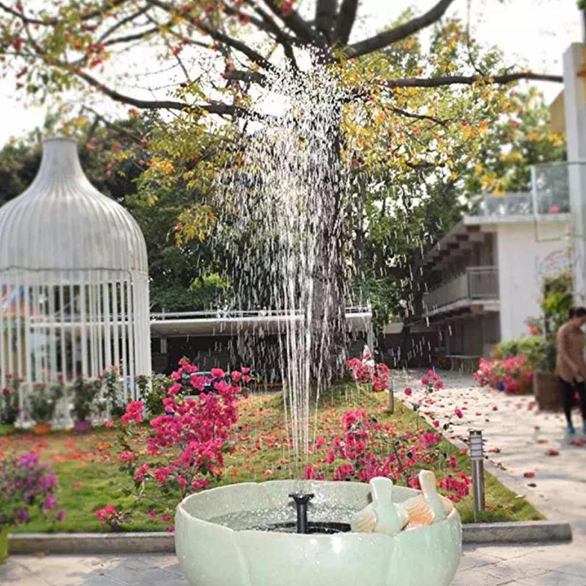 Eine Gartenszene mit einem weißen Vogelkäfig, einem grünen Brunnen, der Wasser sprüht, und leuchtend rosa Blumen auf grünem Gras.
