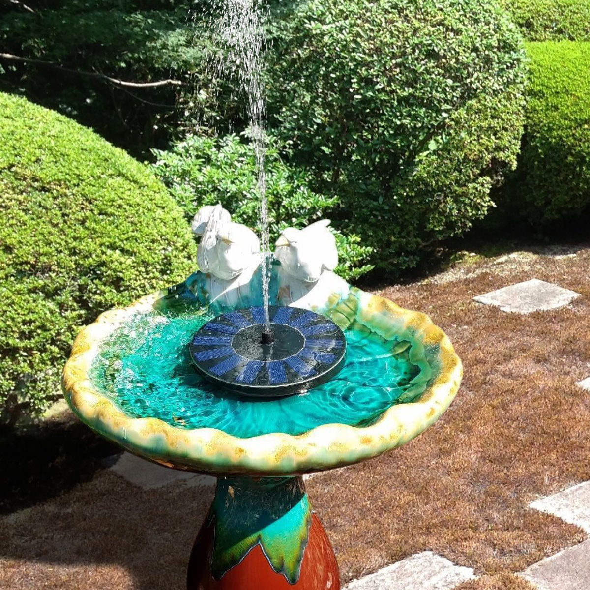 Ein solarbetriebener Vogelbadbrunnen mit zwei weißen Vögeln auf einem Sockel, in einem Garten.