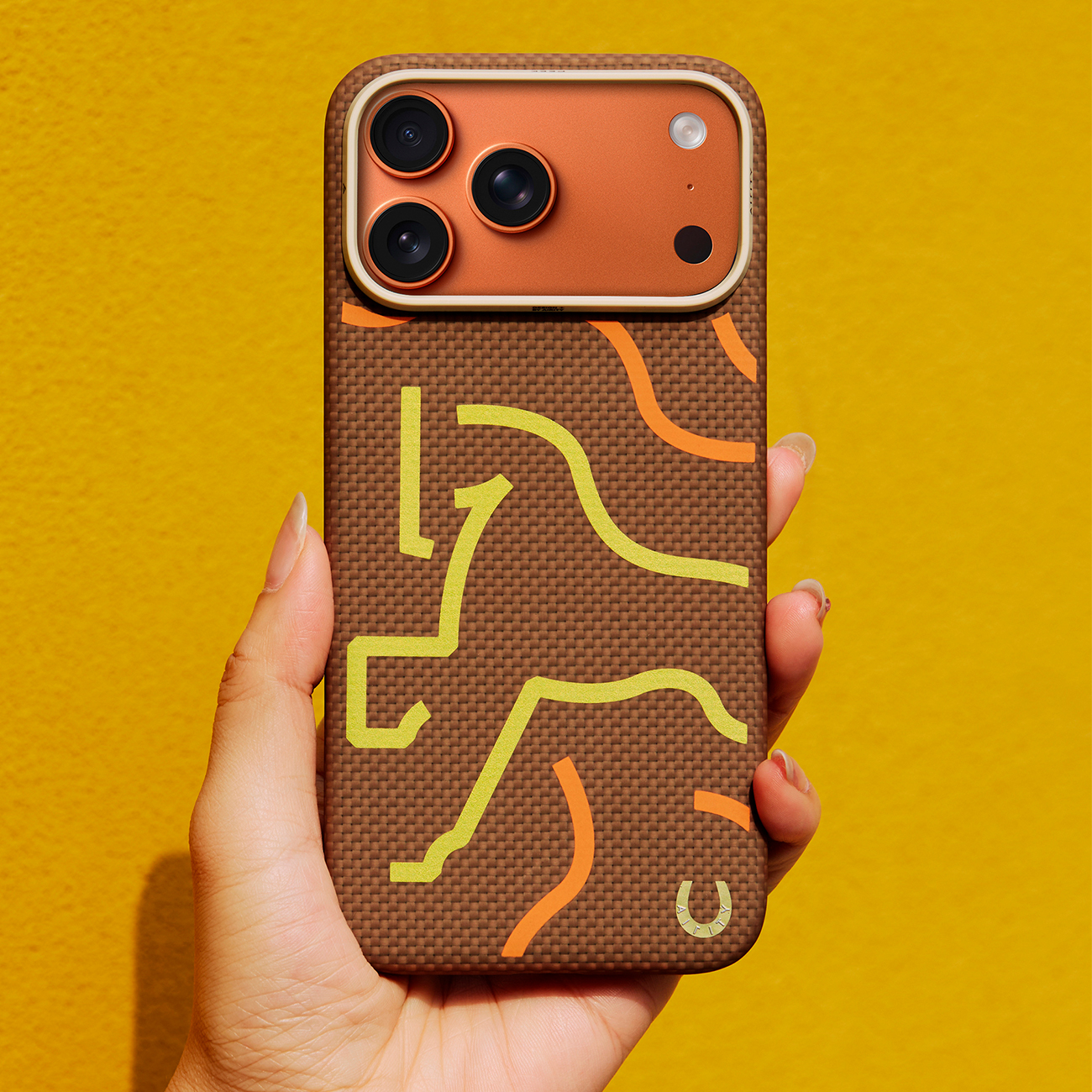 Una mano sostiene una funda de teléfono marrón con líneas abstractas amarillas y naranjas y un logo de herradura.