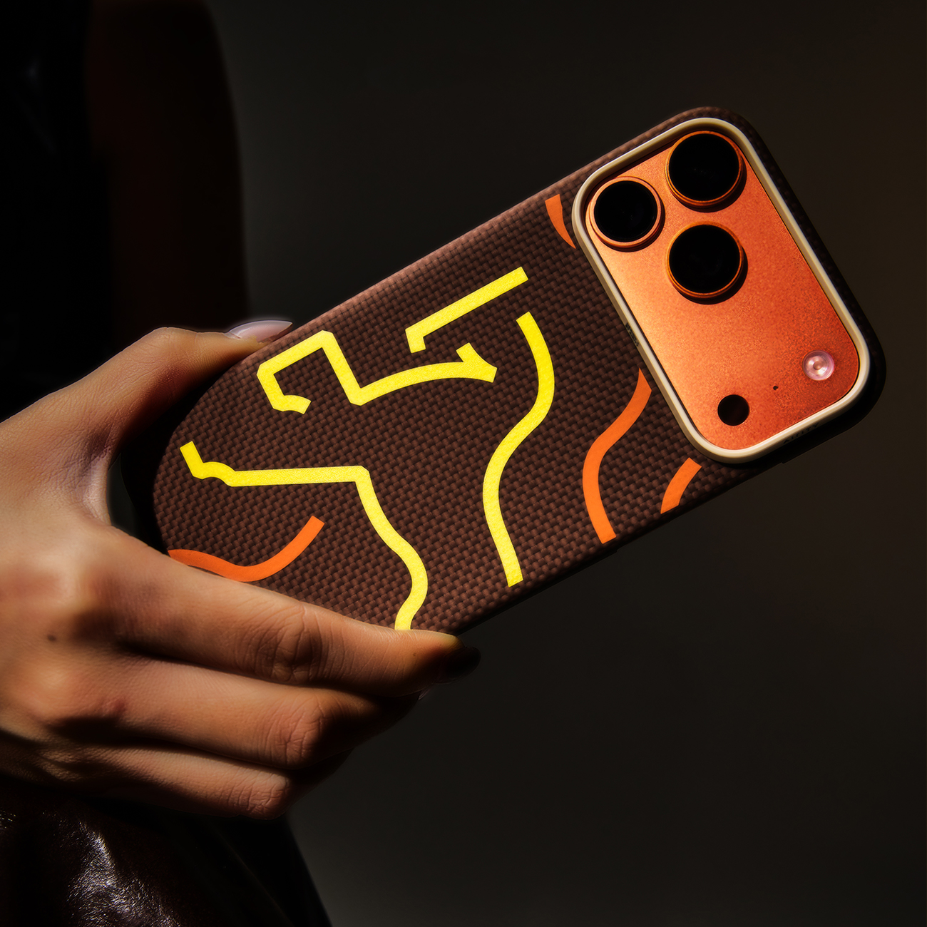 Una mano sostiene una funda de teléfono con textura de fibra de carbono marrón y líneas onduladas amarillas/naranjas.