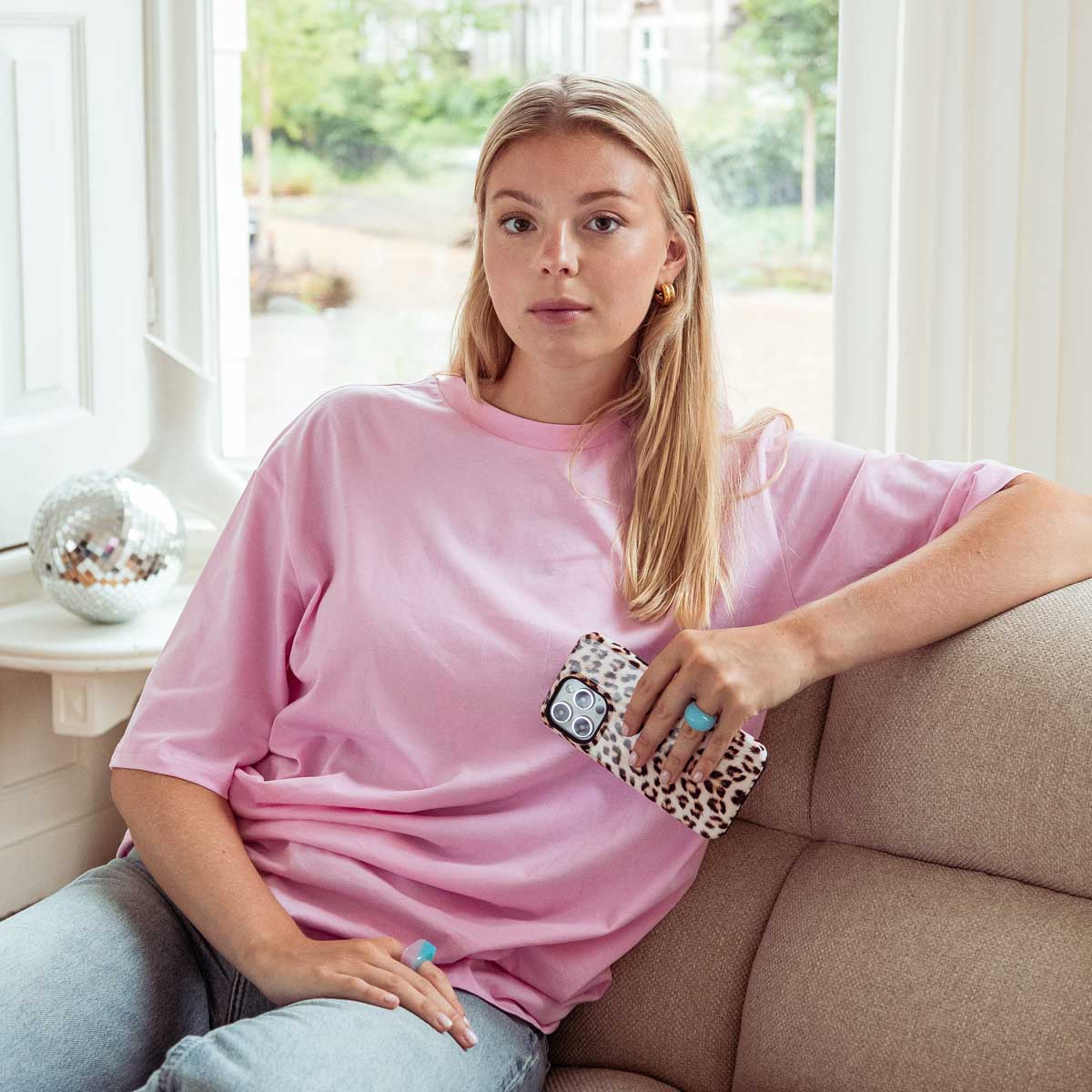 Una mujer con camiseta rosa se sienta en un sofá sosteniendo un teléfono con estampado de leopardo.