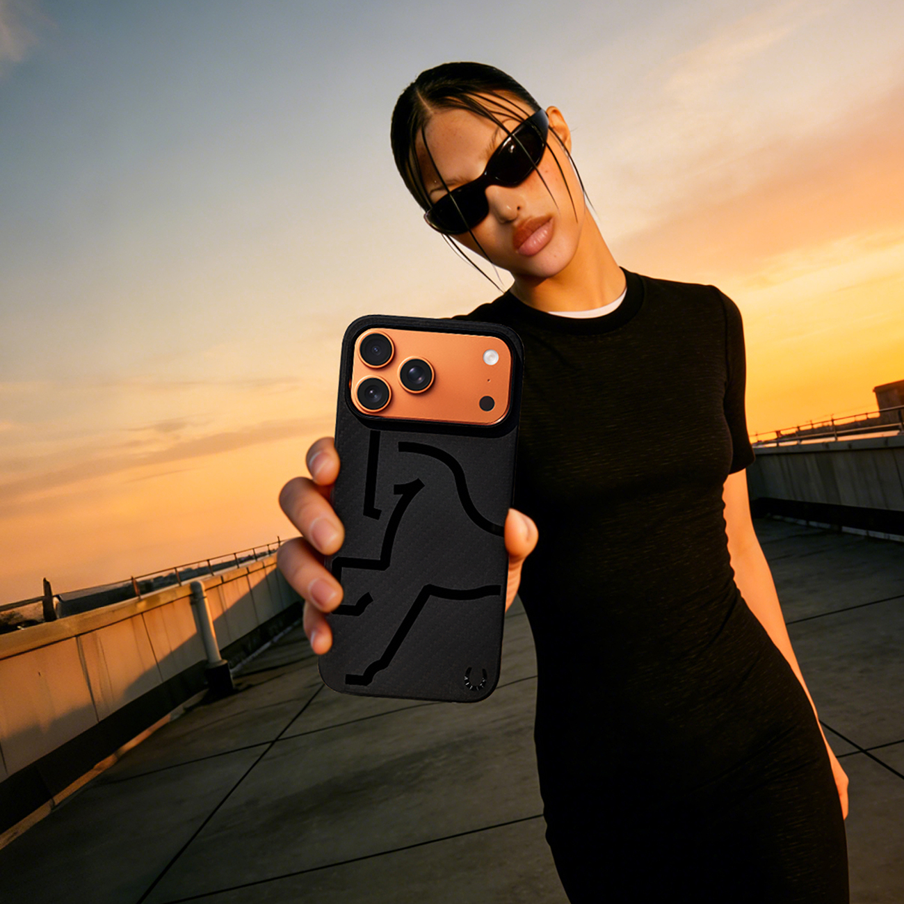 Mujer con gafas de sol sostiene teléfono con funda negra y cámara naranja al atardecer.
