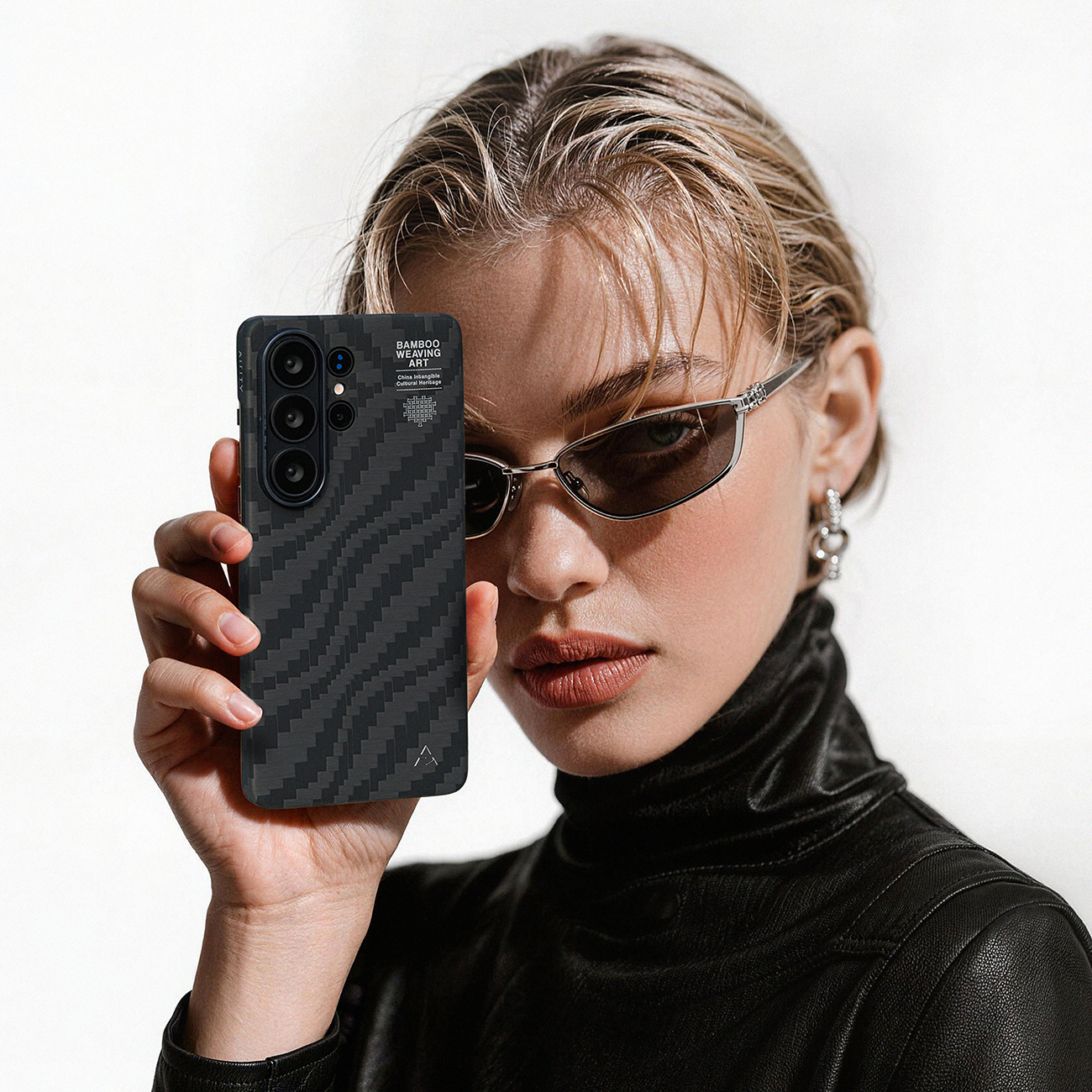 Una mujer con gafas de sol sostiene un teléfono con funda de fibra de carbono frente a su cara.