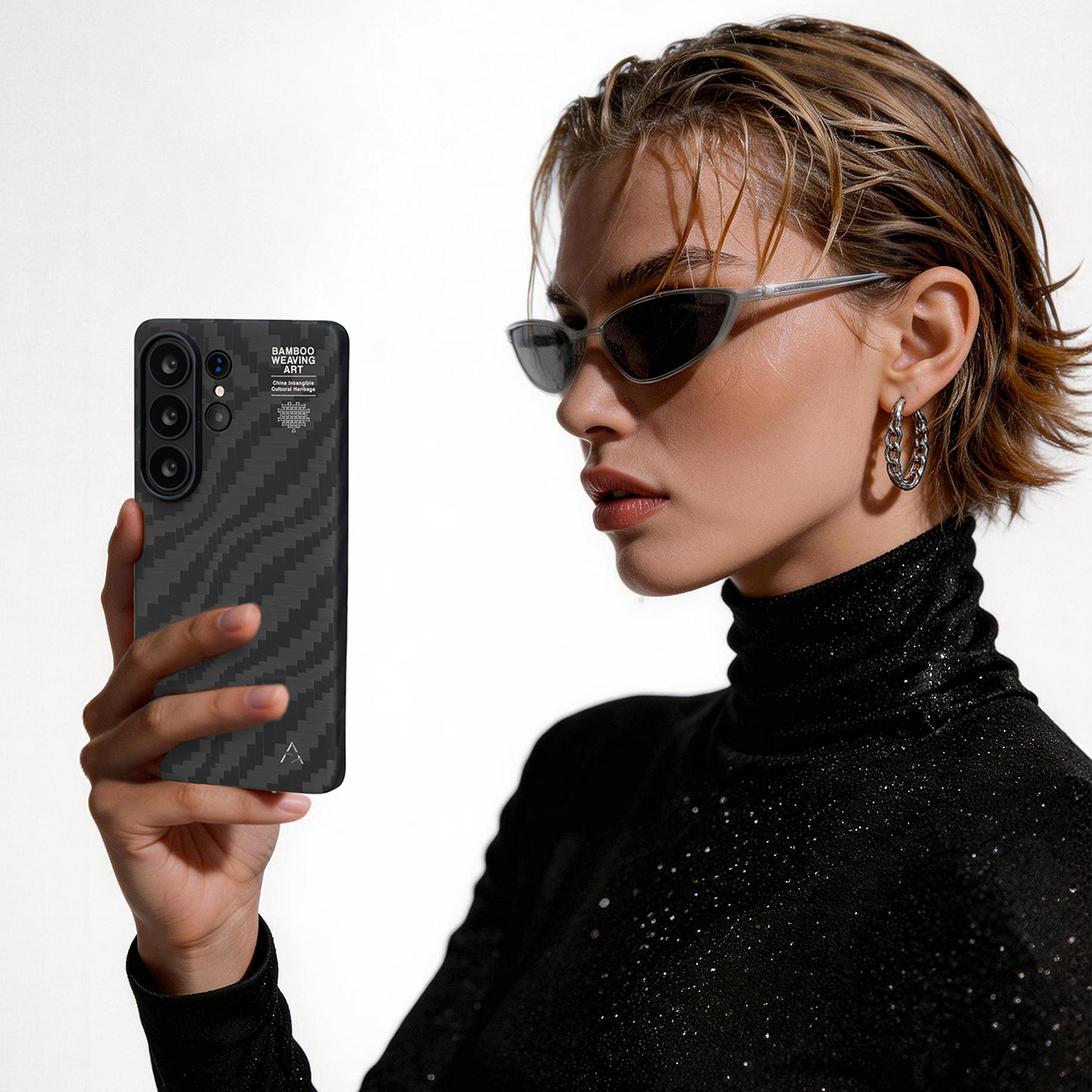 Mujer con gafas de sol sostiene teléfono con funda de carbono. Top negro brillante.