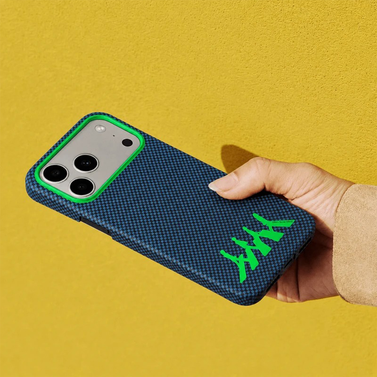 Mano sosteniendo una funda de teléfono azul con ribete verde y logo verde sobre fondo amarillo.