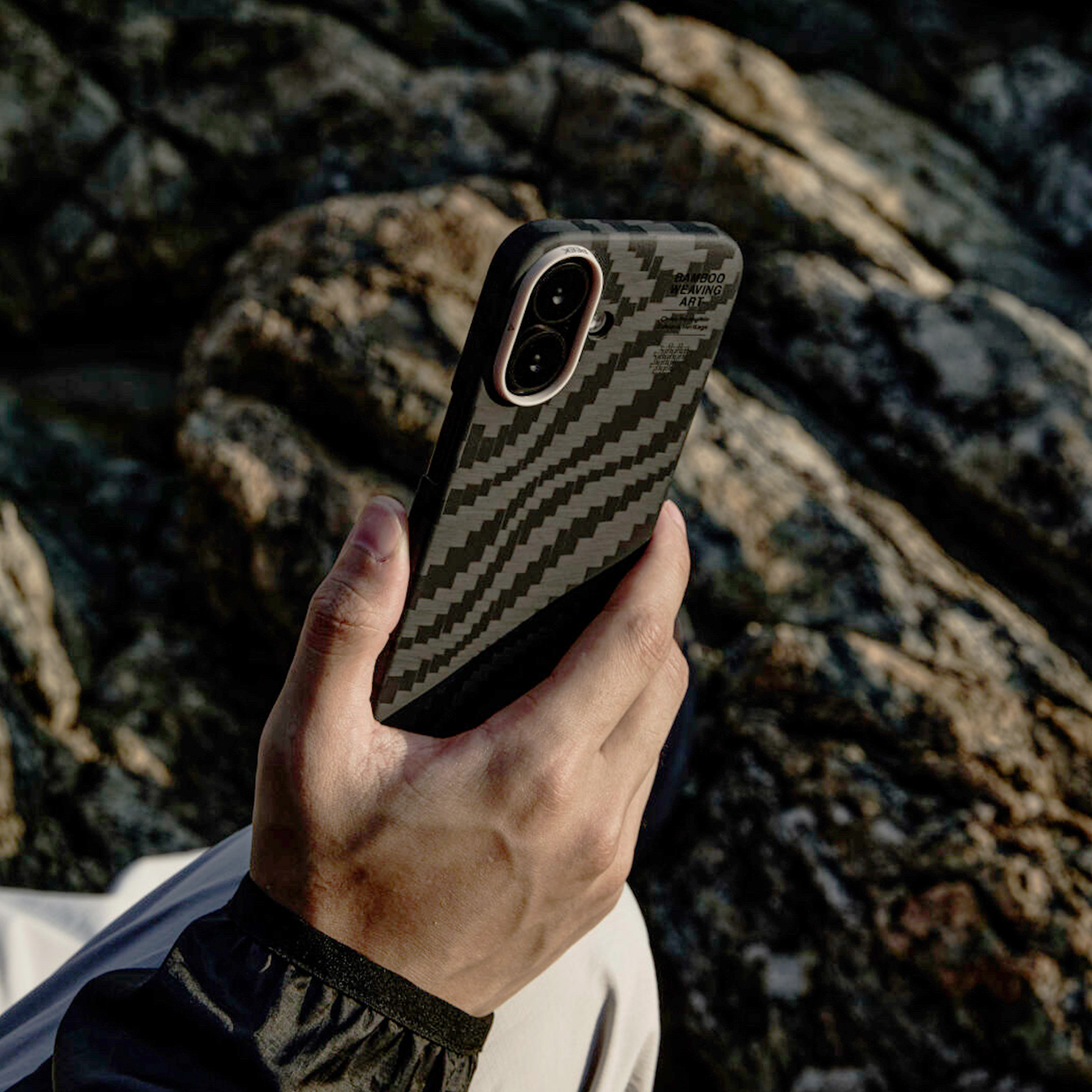 Una mano sostiene un smartphone con funda de patrón de carbono negro frente a rocas.