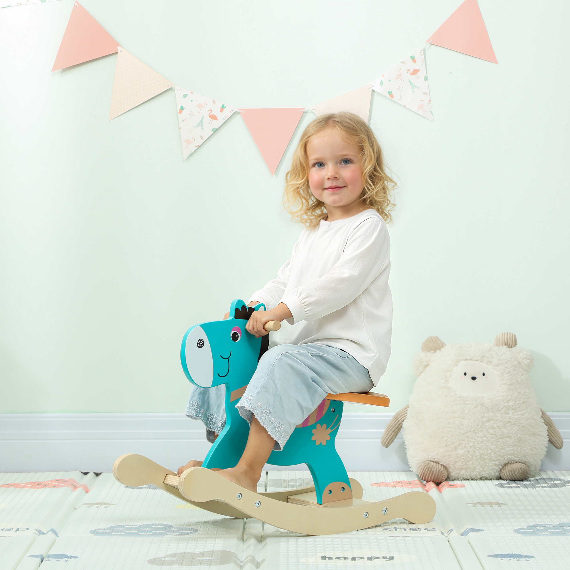 Una bambina bionda cavalca un cavalluccio a dondolo turchese davanti a una parete con ghirlande.