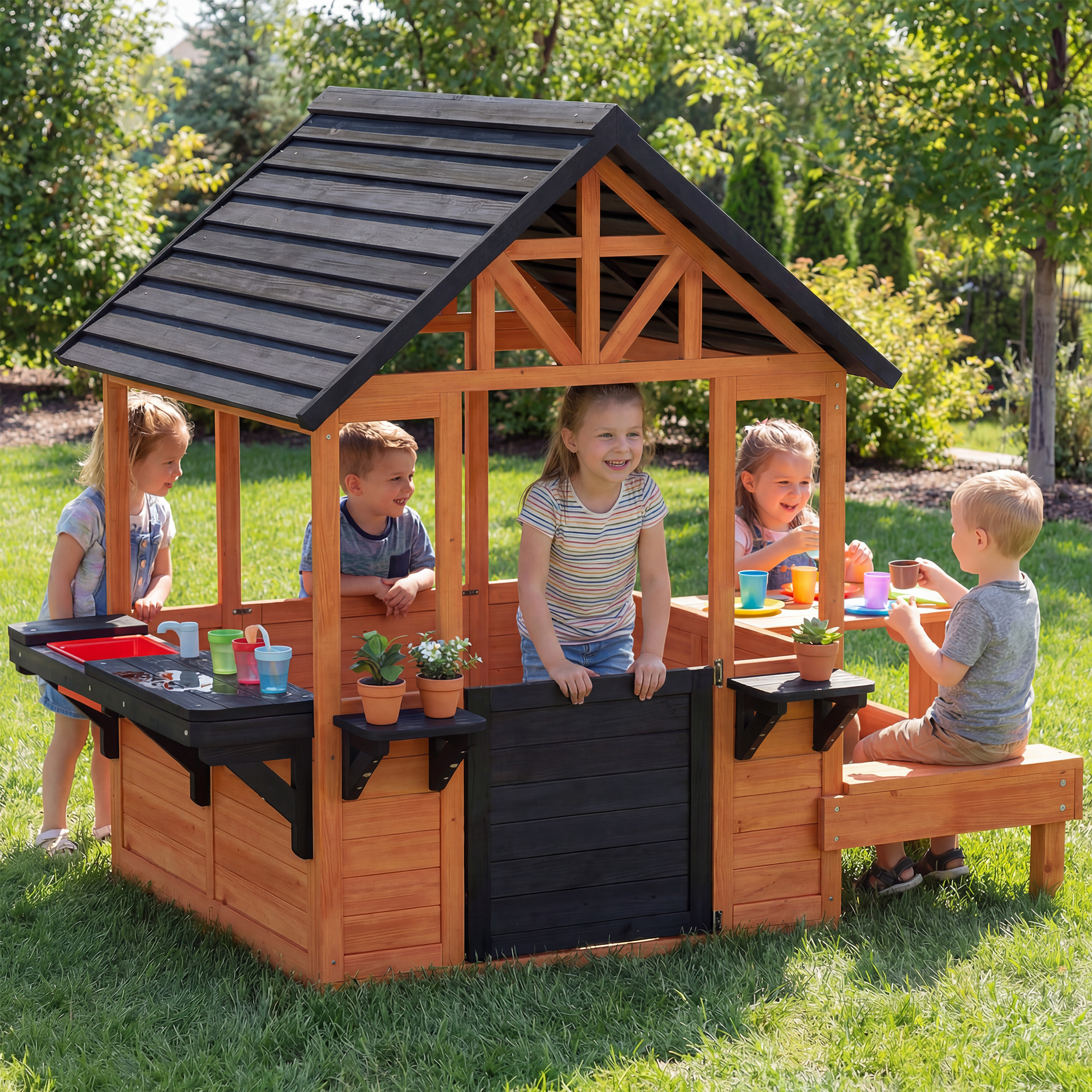 Cinque bambini giocano in una casa giochi di legno con cucina e panca.