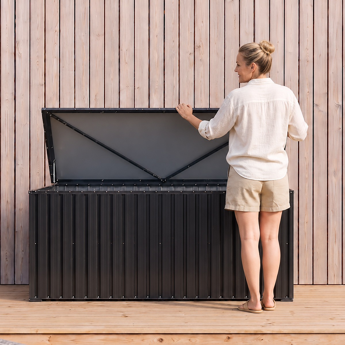 Frau öffnet einen dunklen Aufbewahrungsschrank auf einer Holzterrasse vor Holzwand.