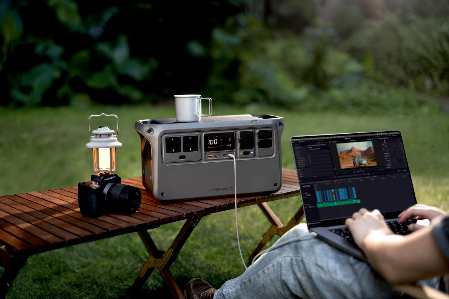 A portable power station sits on a wooden table with a camera, lantern, and mug.