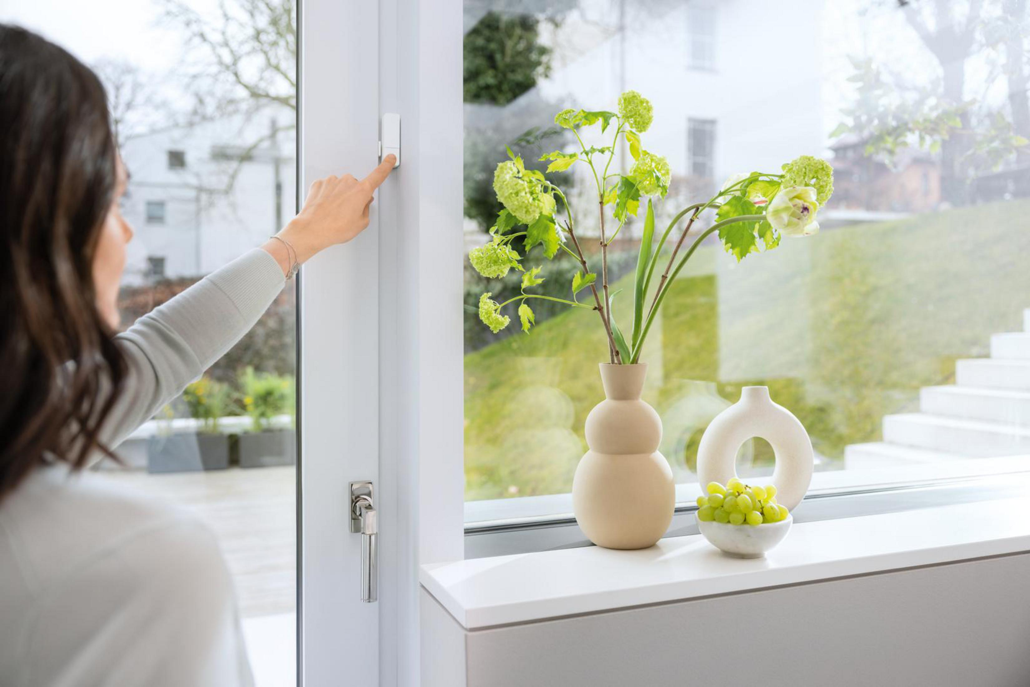 Frau drückt einen weißen Knopf an einem weißen Fensterrahmen neben Blumen.