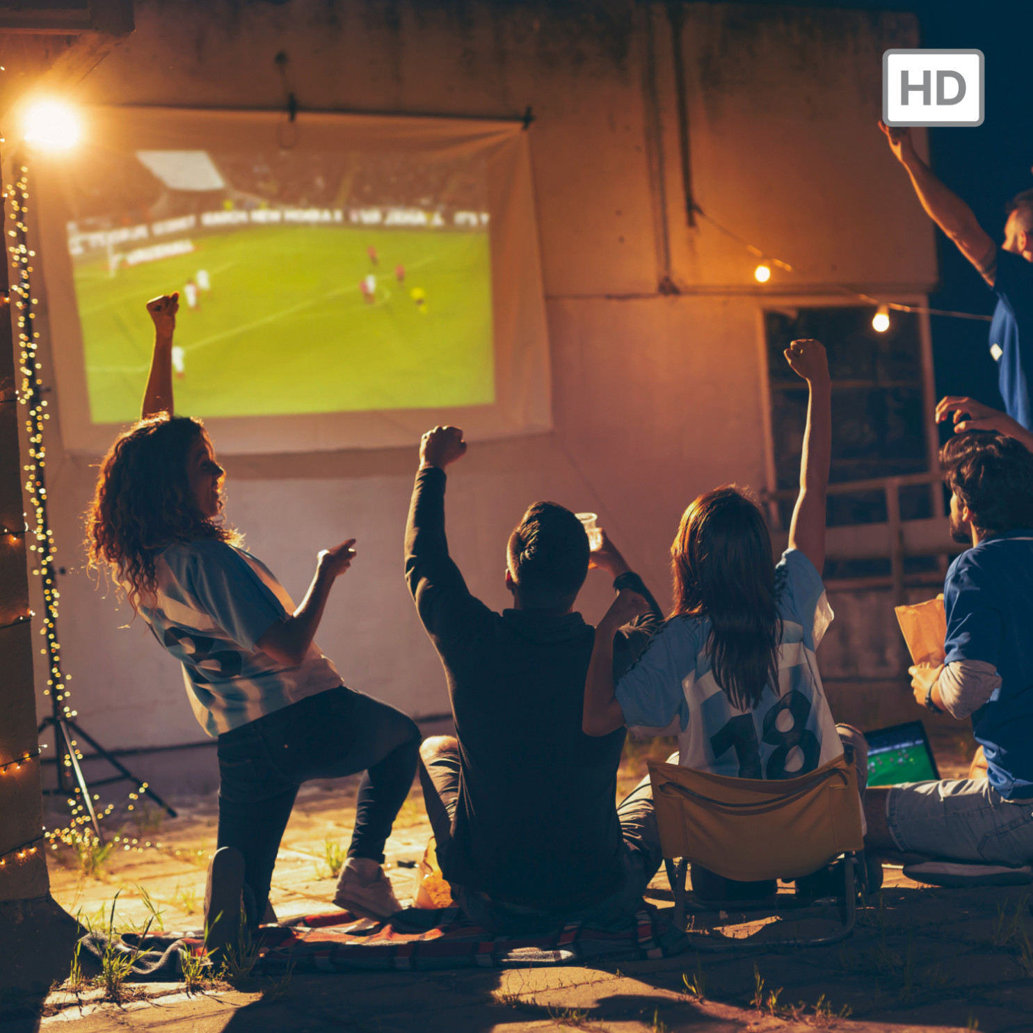 Fans jubeln vor Leinwand mit Fußballspiel bei Nacht, beleuchtet von Lichterketten.