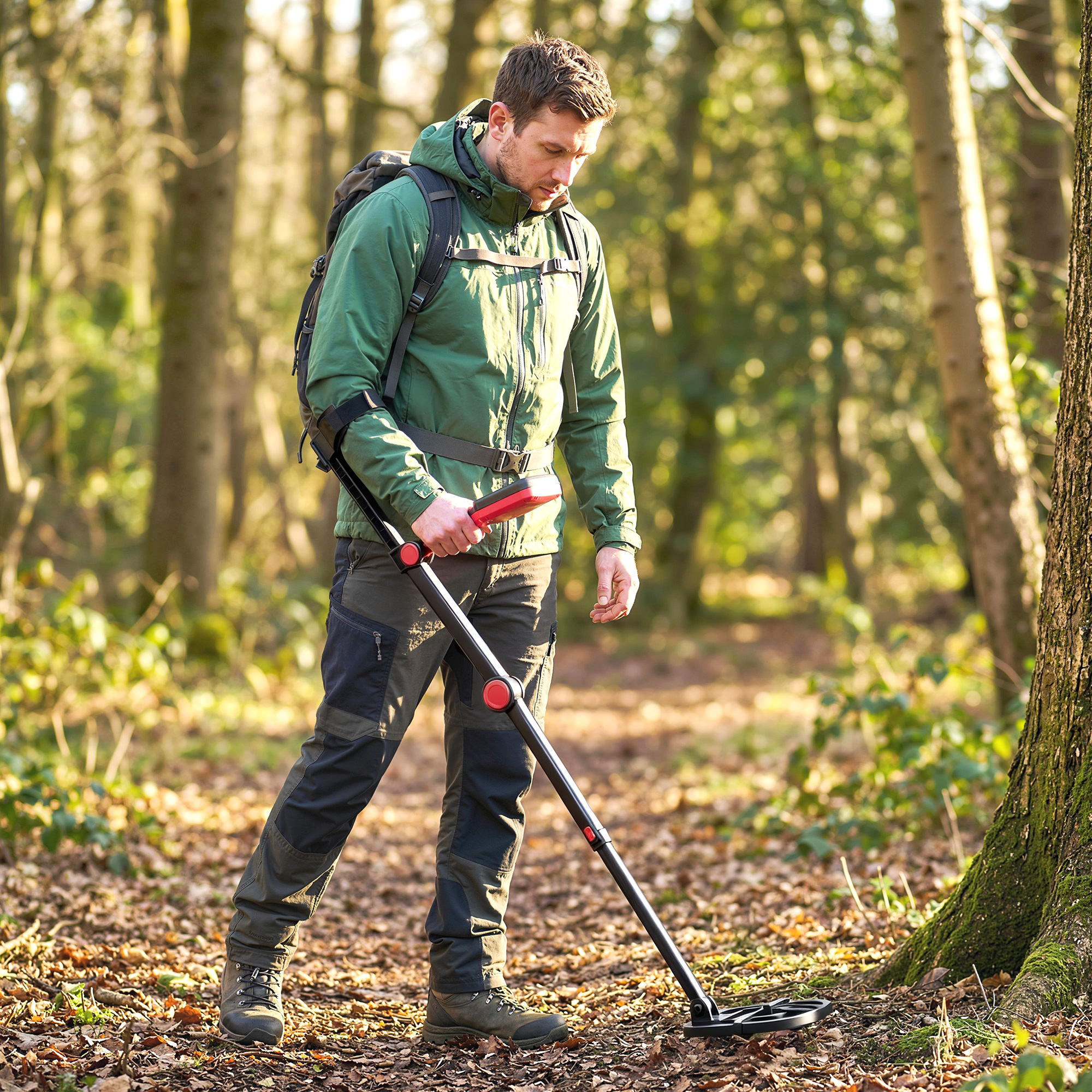 Uomo con metal detector su un sentiero coperto di foglie in un bosco.