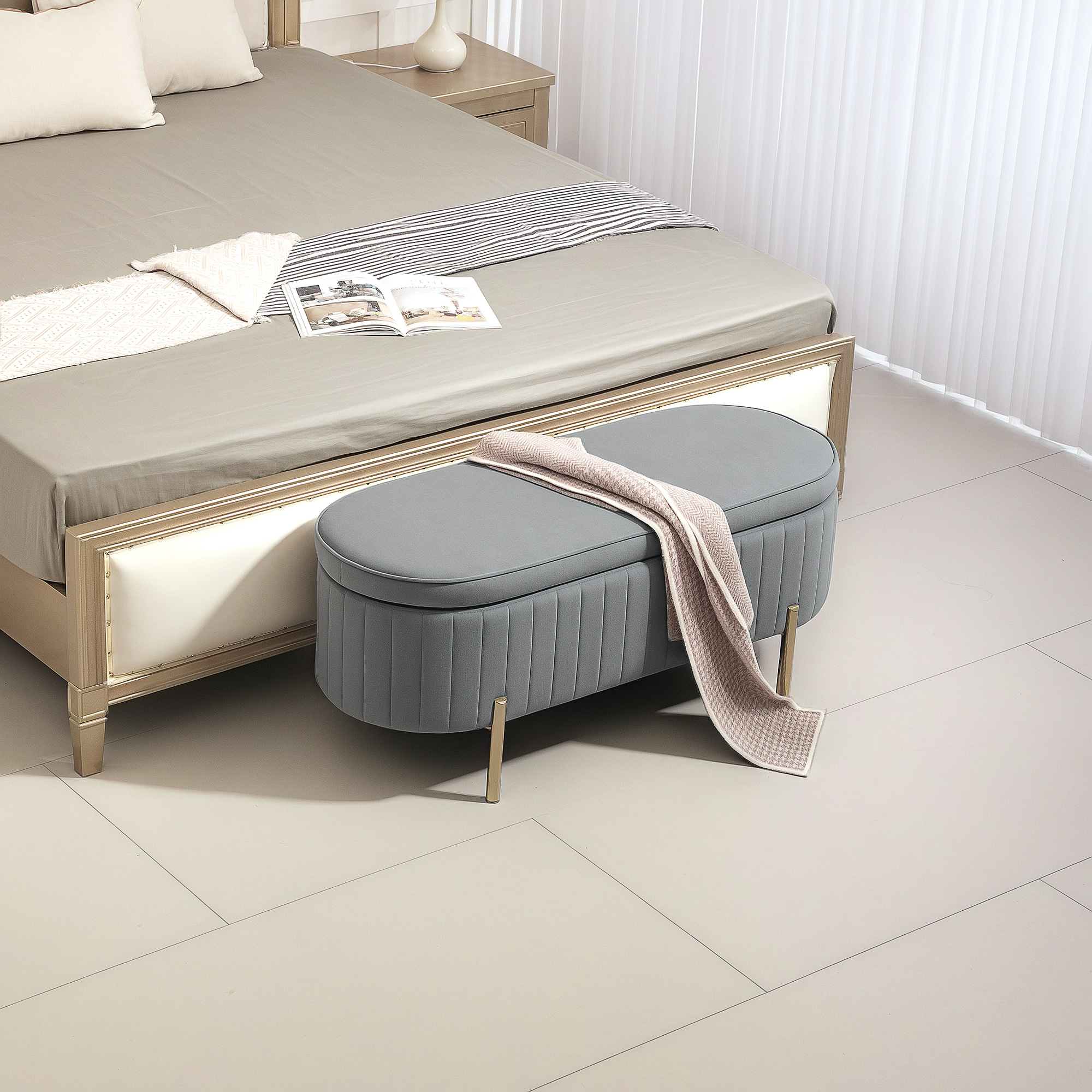 Pouf grigio con coperta davanti a un letto con cuscini e libro. Un comodino con lampada.