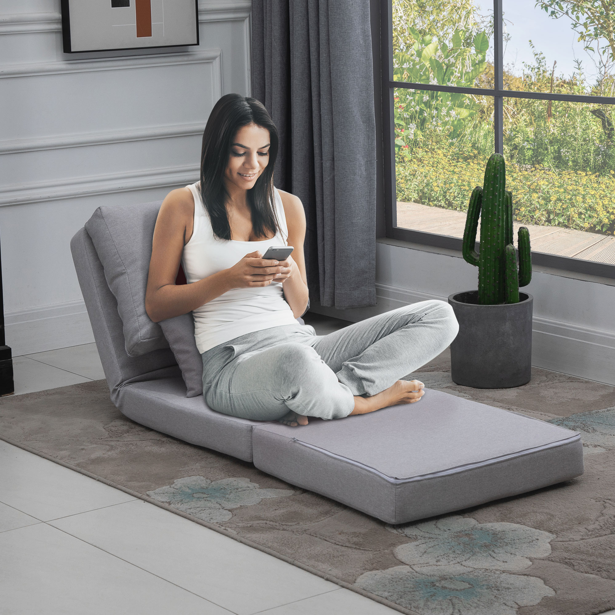 Una donna su un divano letto grigio guarda il telefono. Un cactus è vicino alla finestra.