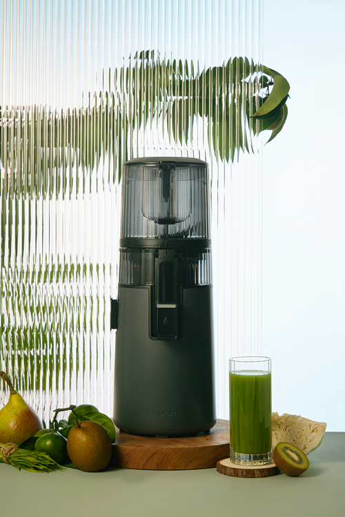 Dunkelgrüner Entsafter mit Früchten und Glas Saft vor geriffeltem Hintergrund.