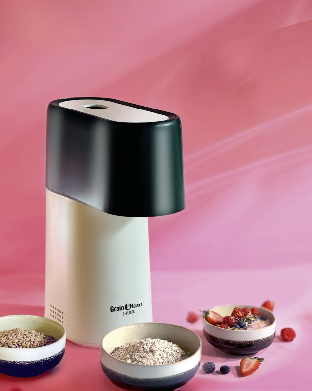 Ein Müsli-Flocker steht neben Schalen mit Getreide und Haferflocken. Ein Schälchen mit Müsli und Beeren steht rechts.