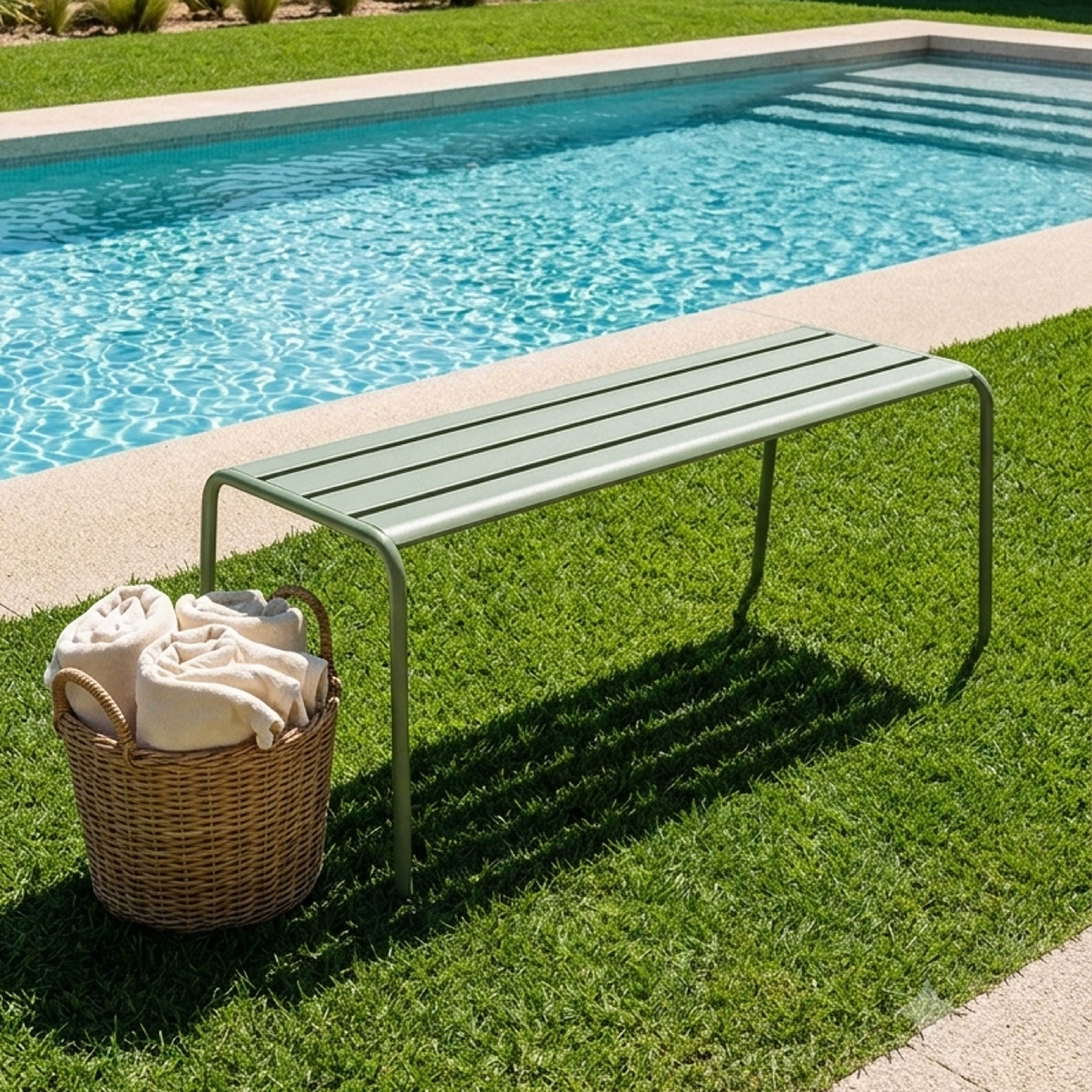 Ein grünes Metall-Sitzbank steht auf Gras neben einem Pool. Ein Korb mit Handtüchern.