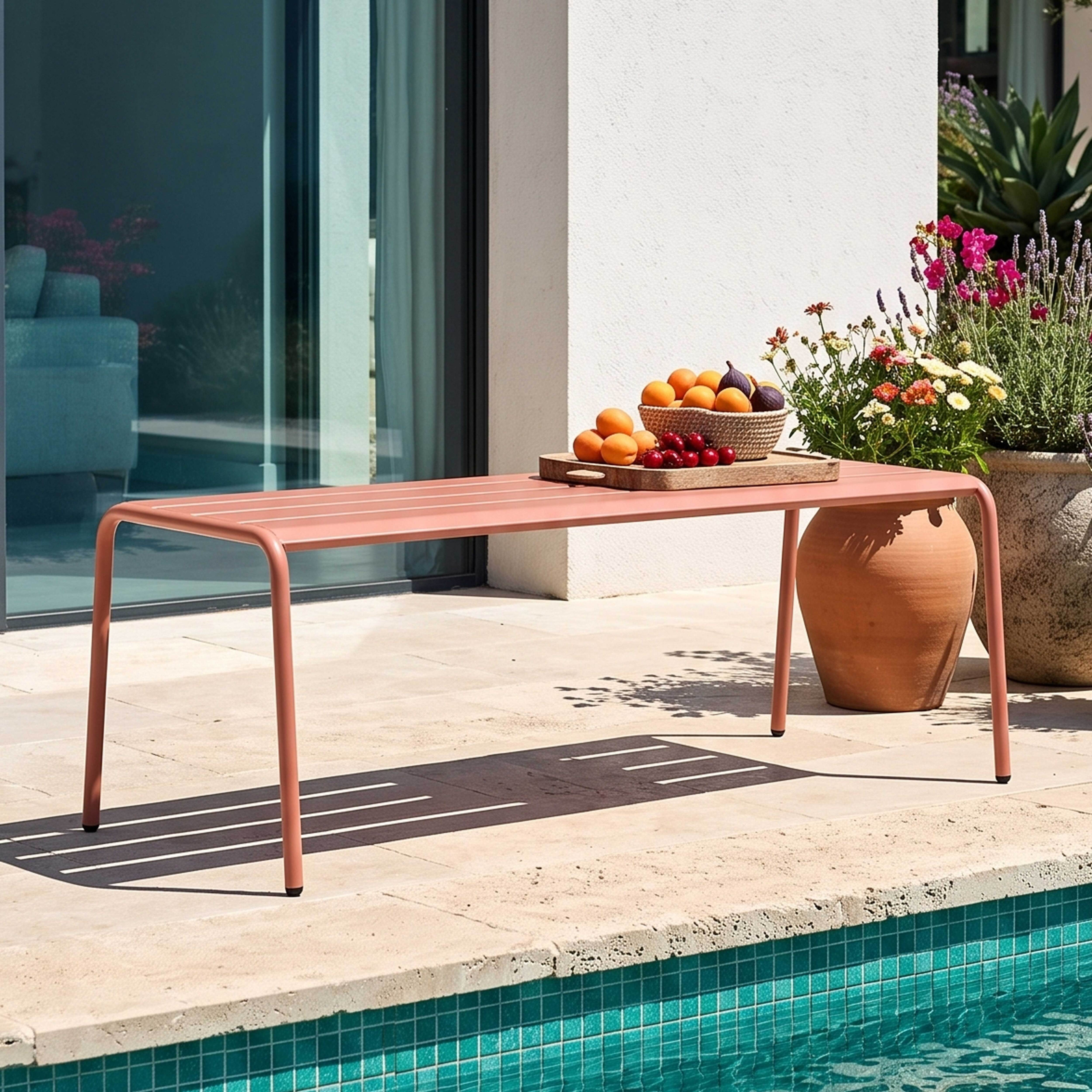 Ein korallfarbener Tisch mit Obst steht an einem Pool. Blumen und große Töpfe sind in der Nähe.