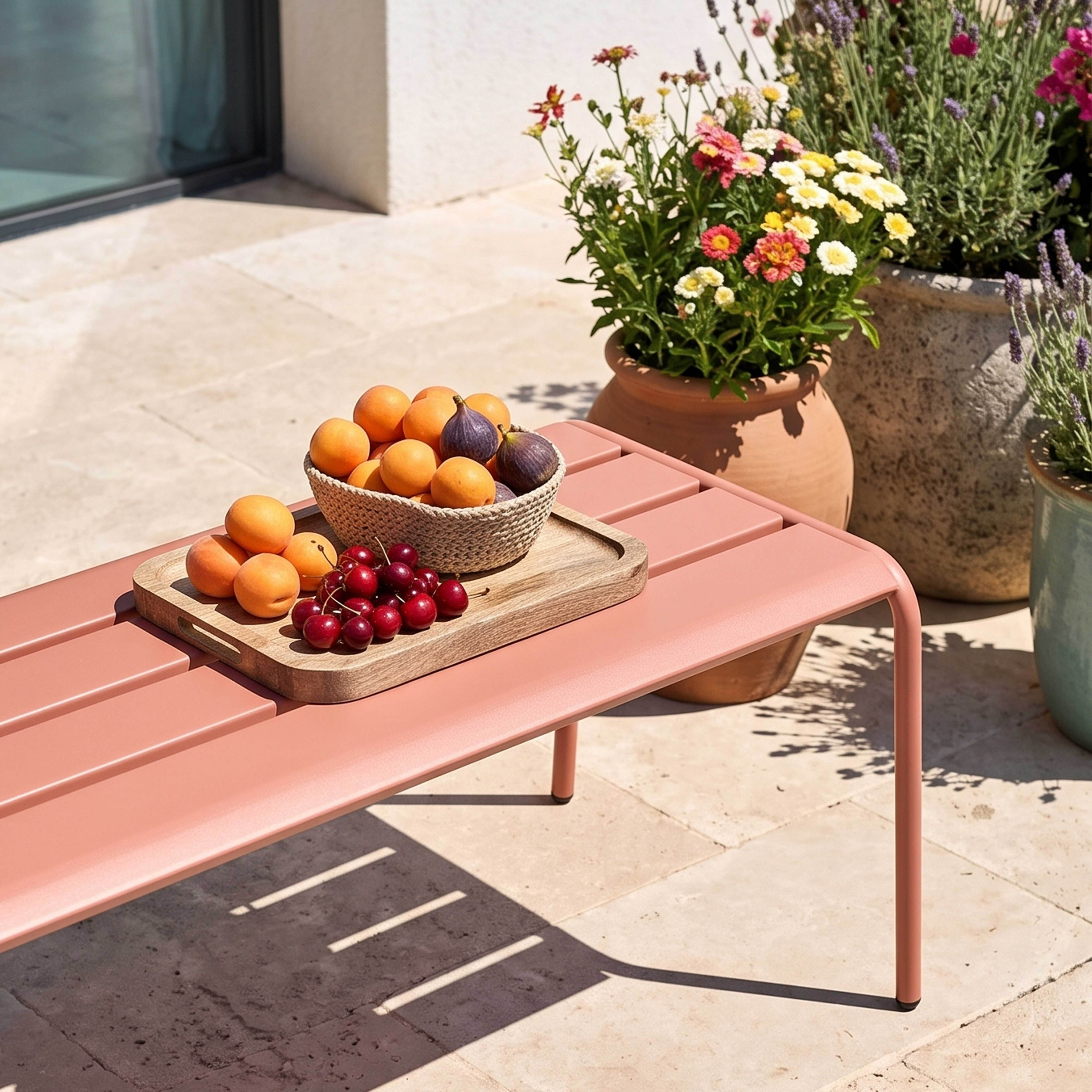 Ein rosa Tisch mit Obstschale, Aprikosen, Feigen und Kirschen, daneben blühende Pflanzen.