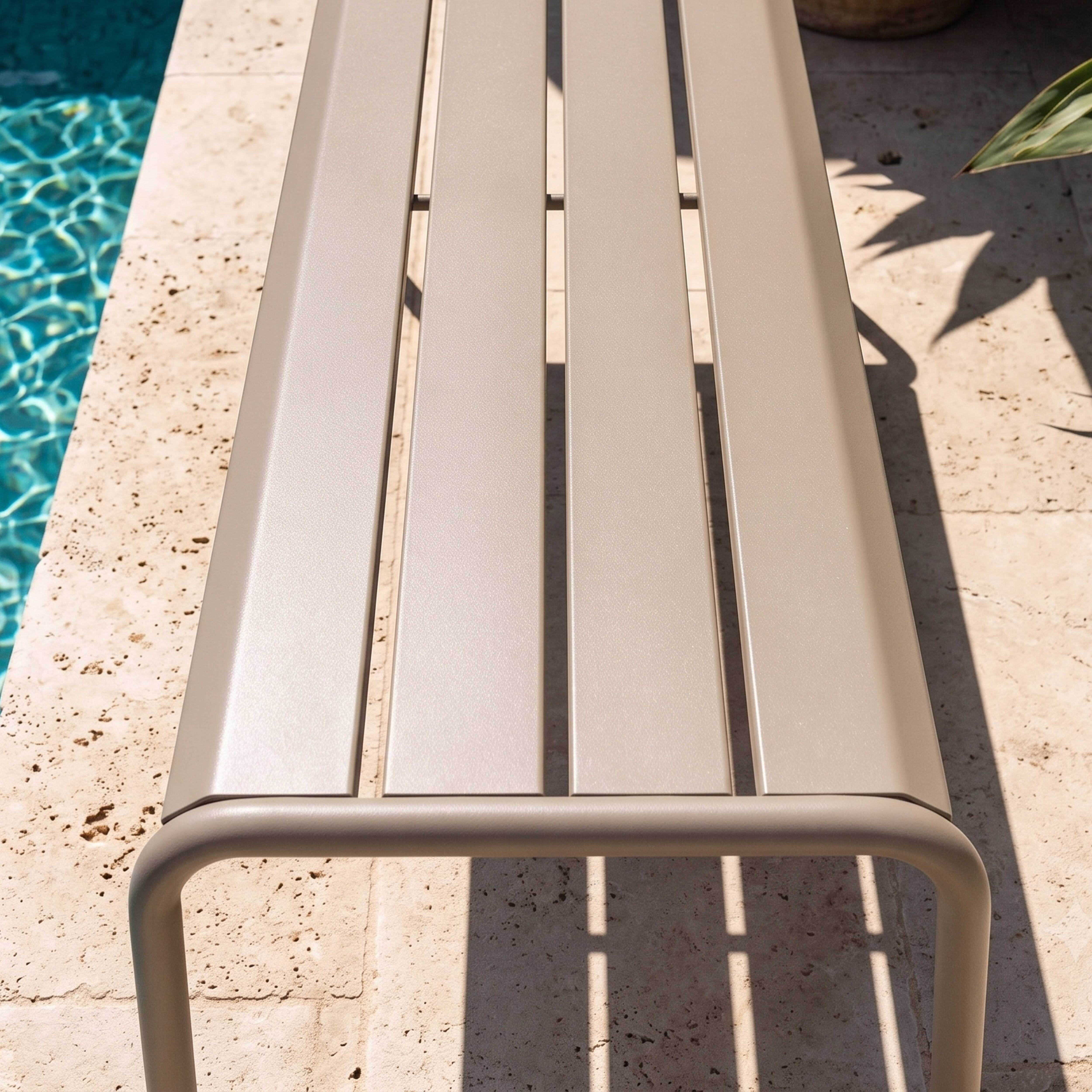 Eine beige Gartenbank steht auf Travertinpflaster neben einem Pool mit blauem Wasser.