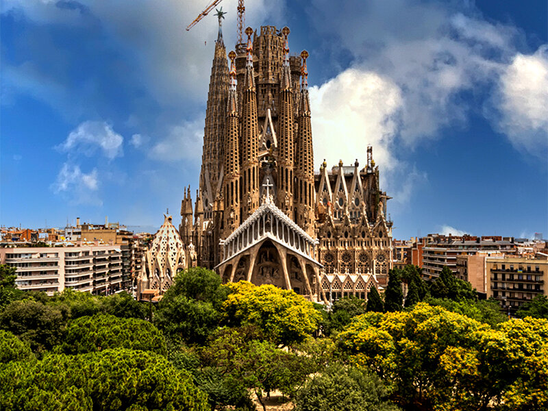 Sagrada Familia en Barcelona bajo un cielo azul con nubes, rodeada de árboles.