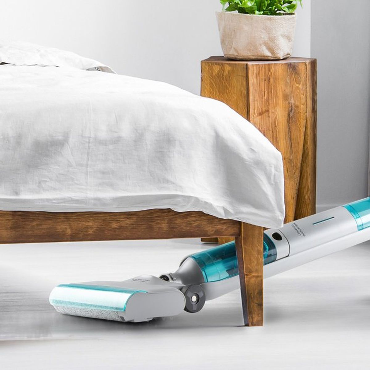 Un aspirateur sous un lit avec des draps blancs et une table de chevet en bois.