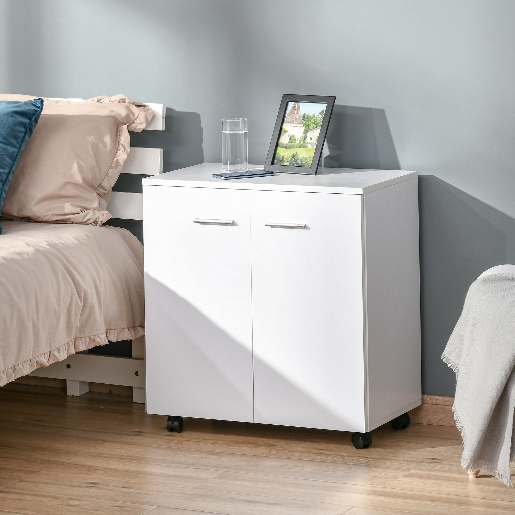Un comodino bianco con due ante, ruote e maniglie argentate accanto a un letto.
