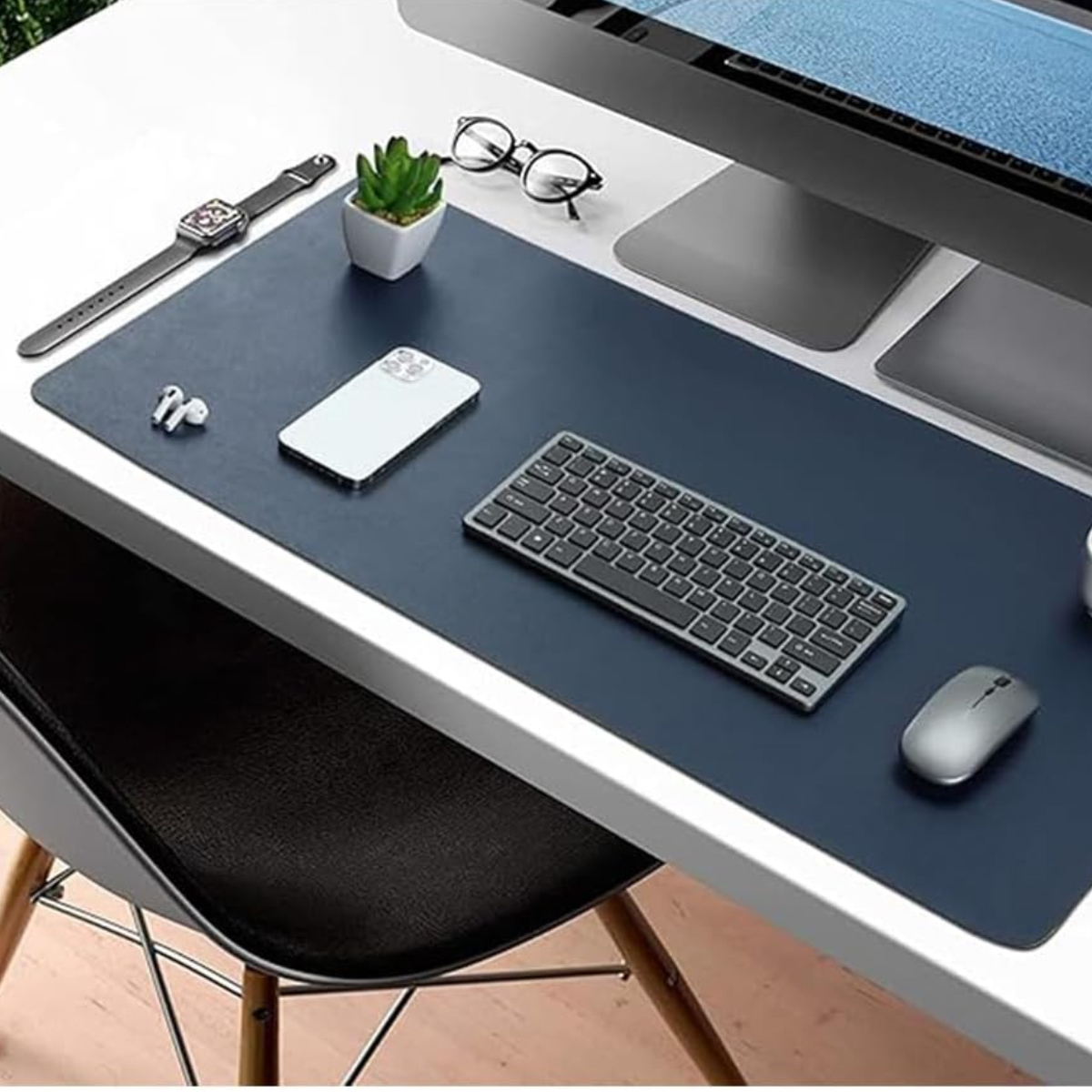 Ein Schreibtisch mit dunkler Unterlage, darauf ein Computer, Tastatur, Maus, Handy, Uhr und Brille.